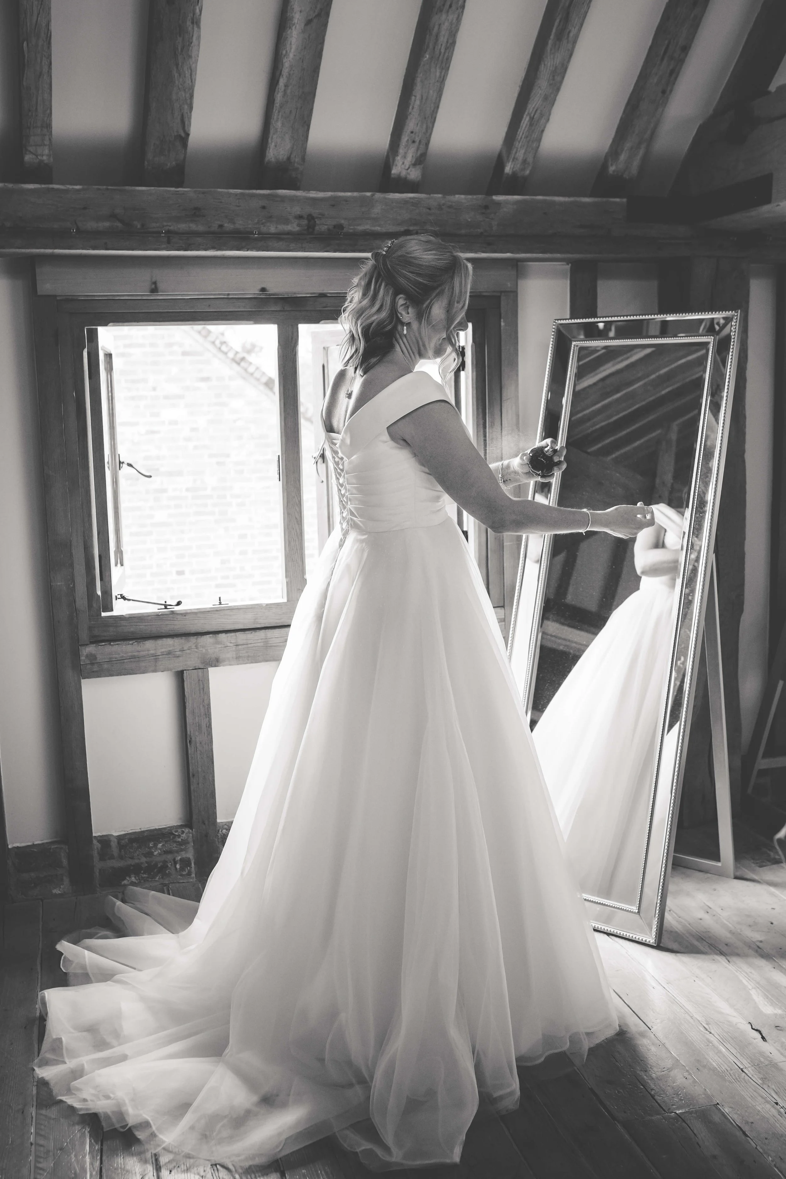 A bride in a wedding dress looks at herself in a full-length mirror in a rustic room with wooden beams and window.