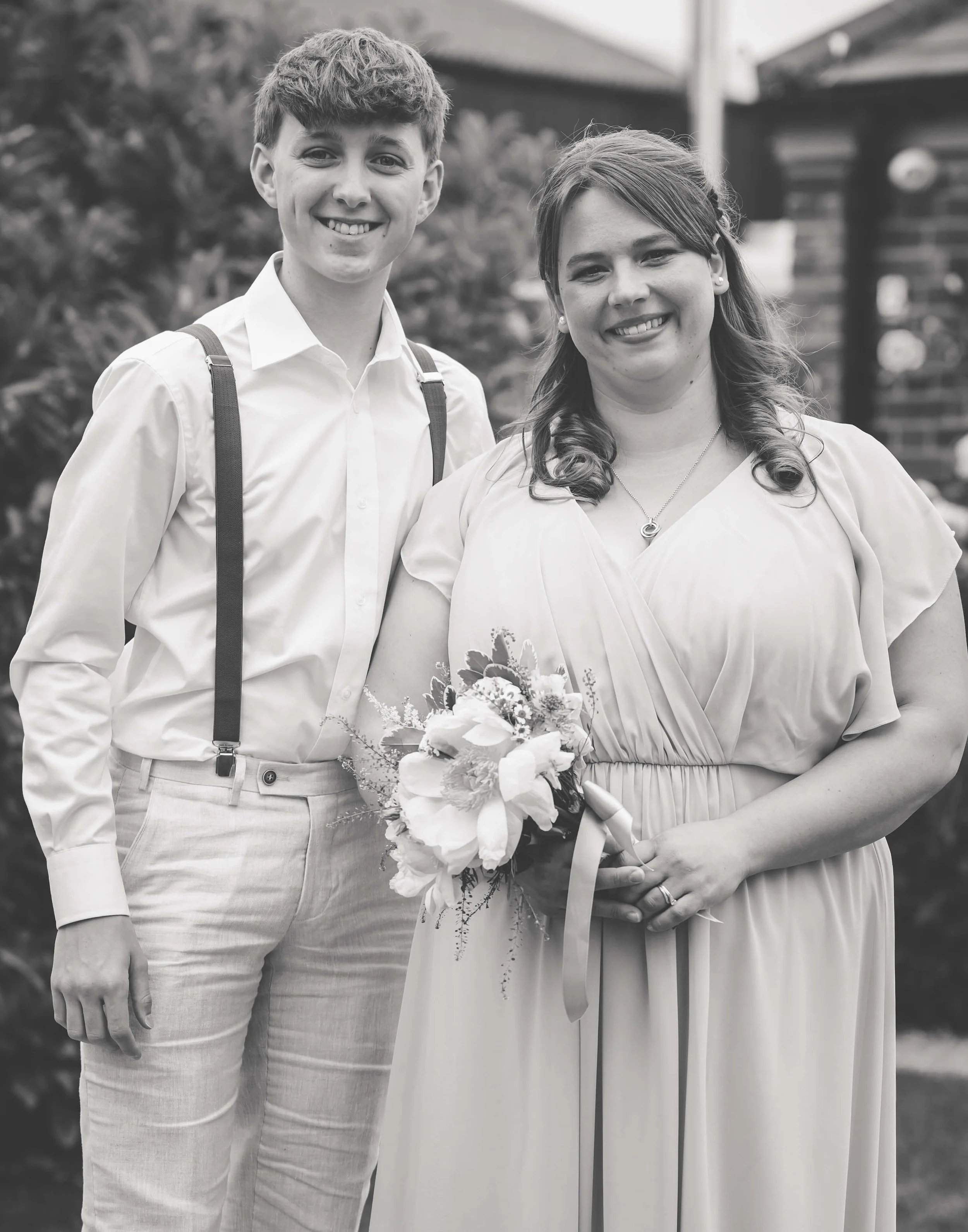 A smiling young man in a white shirt with suspenders standing next to a smiling woman in a light-colored dress holding a bouquet of flowers, outdoors in front of greenery and a brick house.