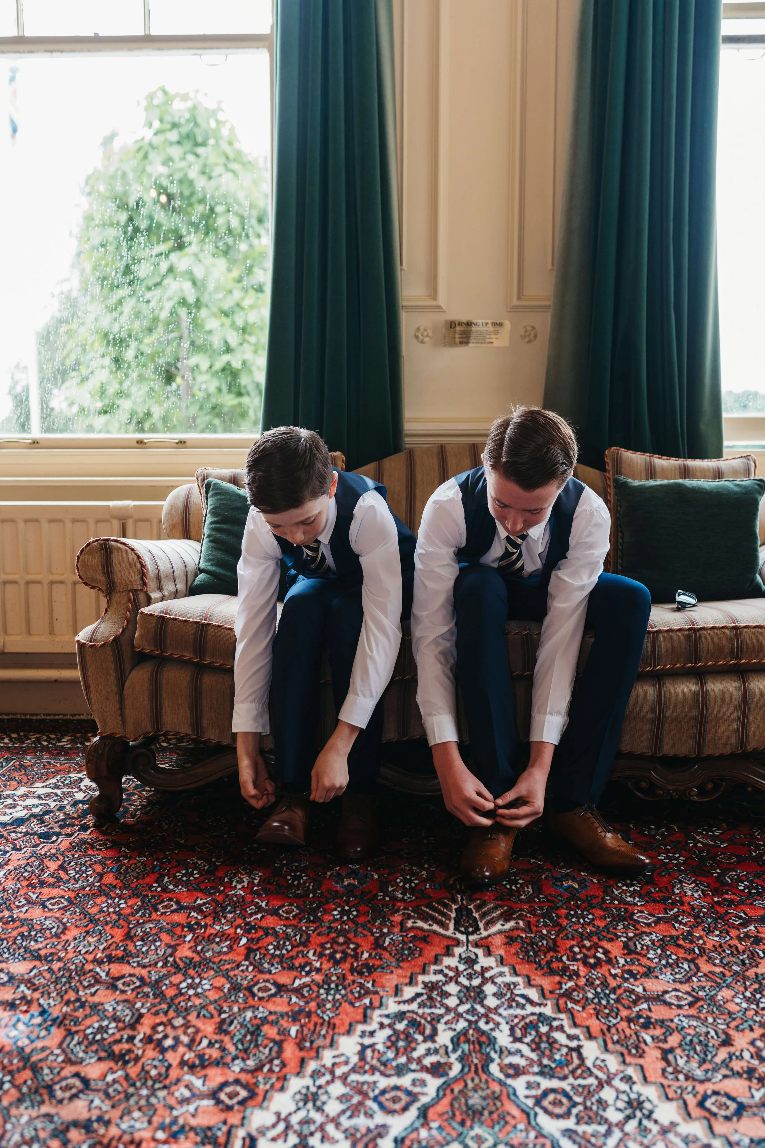 Two boys in formal suits sitting on a vintage sofa, tying their shoelaces, in a room with a large window and green curtains, and a patterned rug on the floor.