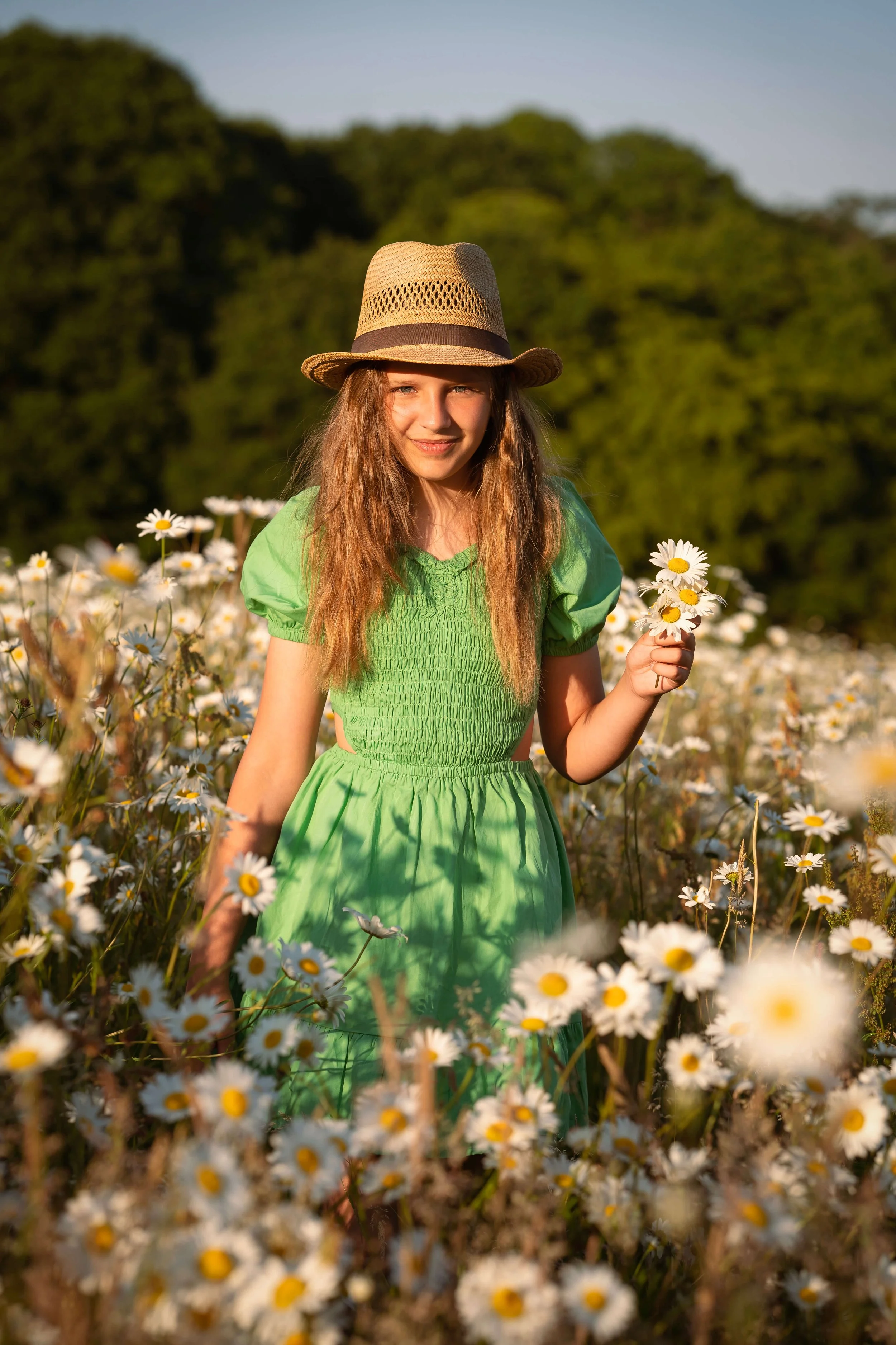 A girl in a green dress and straw hat standing in a field of daisies during sunset.