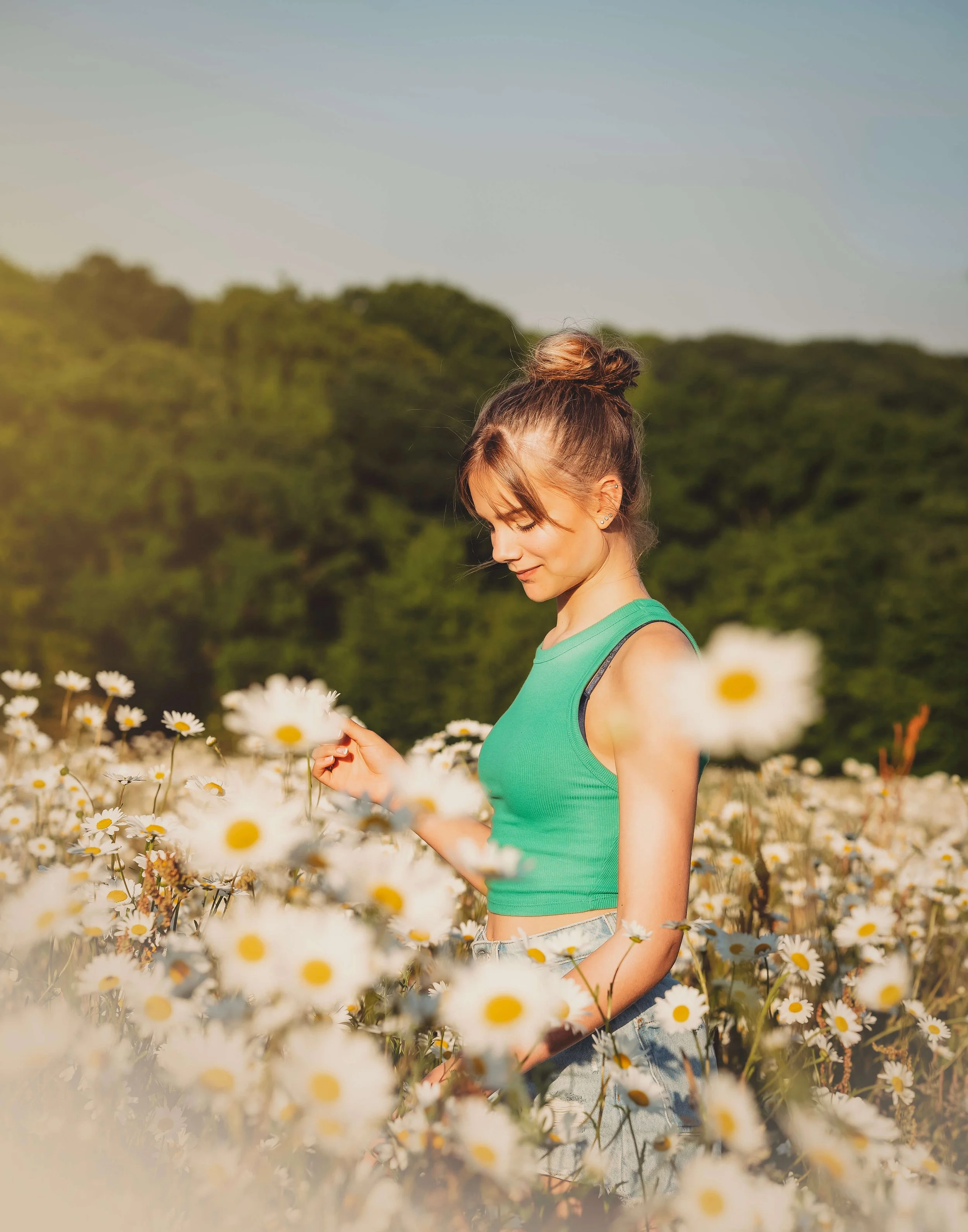 A young woman in a green crop top stands in a field of white daisies with a forested background under a blue sky.