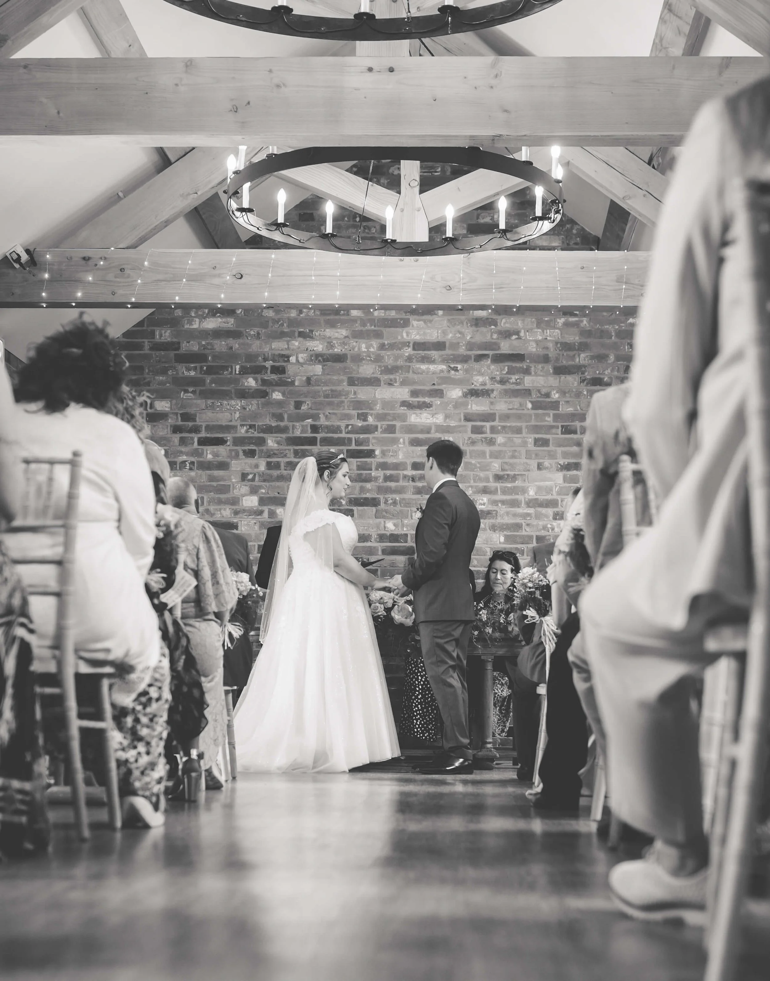 A black-and-white photo of a wedding ceremony with a bride and groom holding hands, facing each other, in a rustic venue with a brick wall backdrop and wooden ceiling beams. Guests are seated on either side, and a woman with a floral dress is sitting