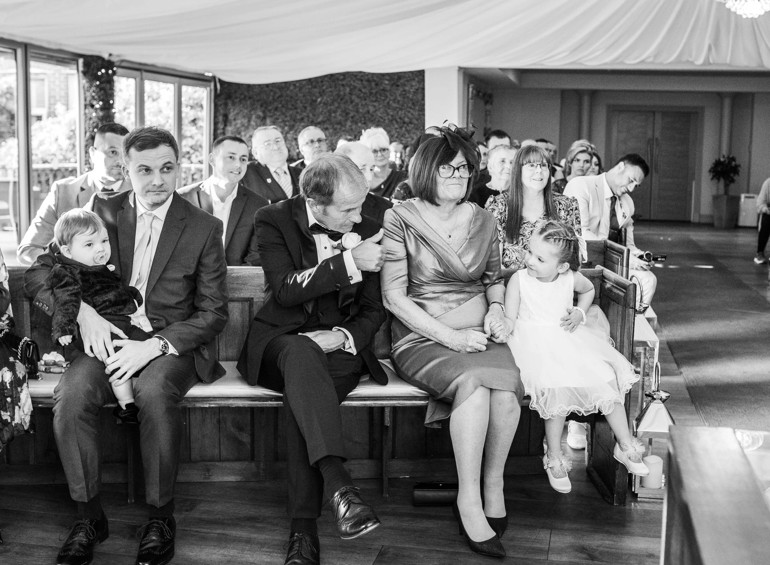 Black and white photo of wedding guests sitting on wooden pews inside a decorated venue. A woman in a shiny dress and glasses is holding the hand of a young girl in a white dress sitting beside her. Men and women in formal attire are seated behind th