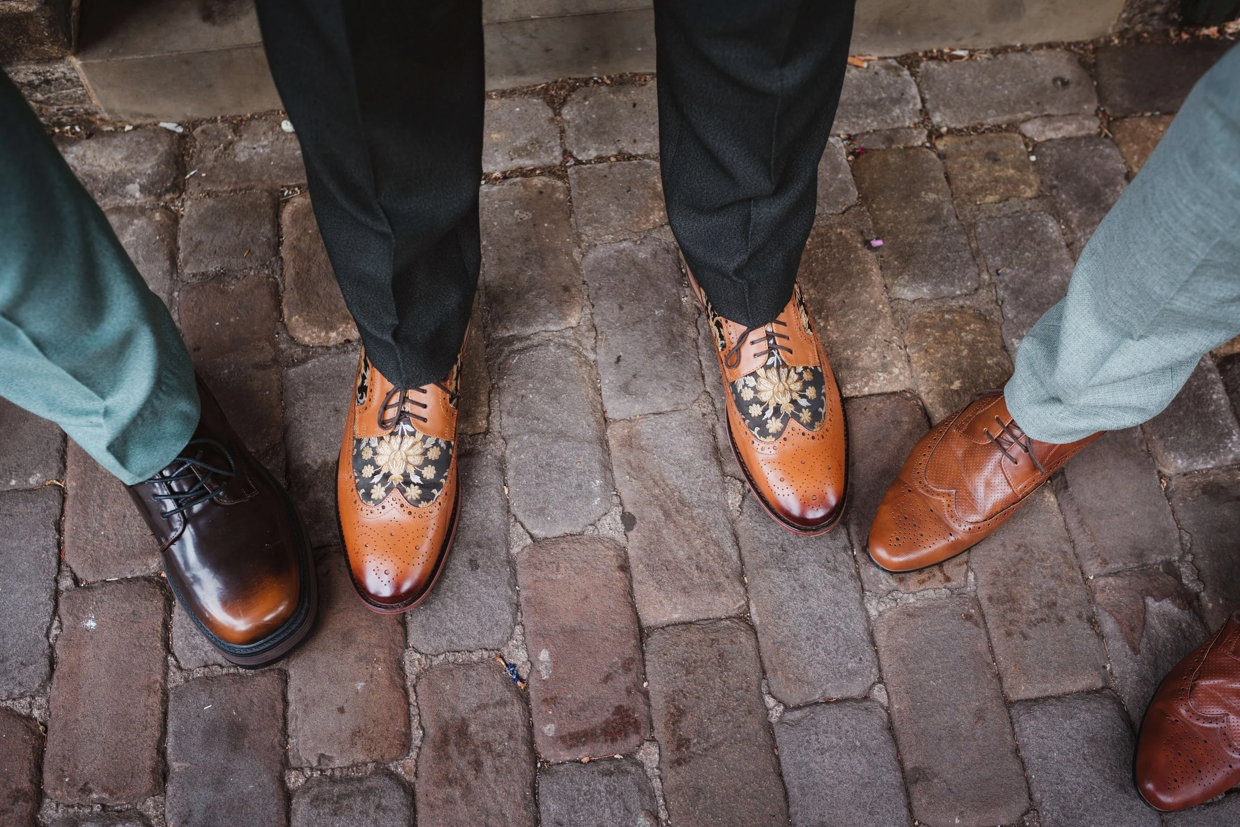 Close-up of three pairs of men's dress shoes on a cobblestone street, with the men wearing different colored suits.