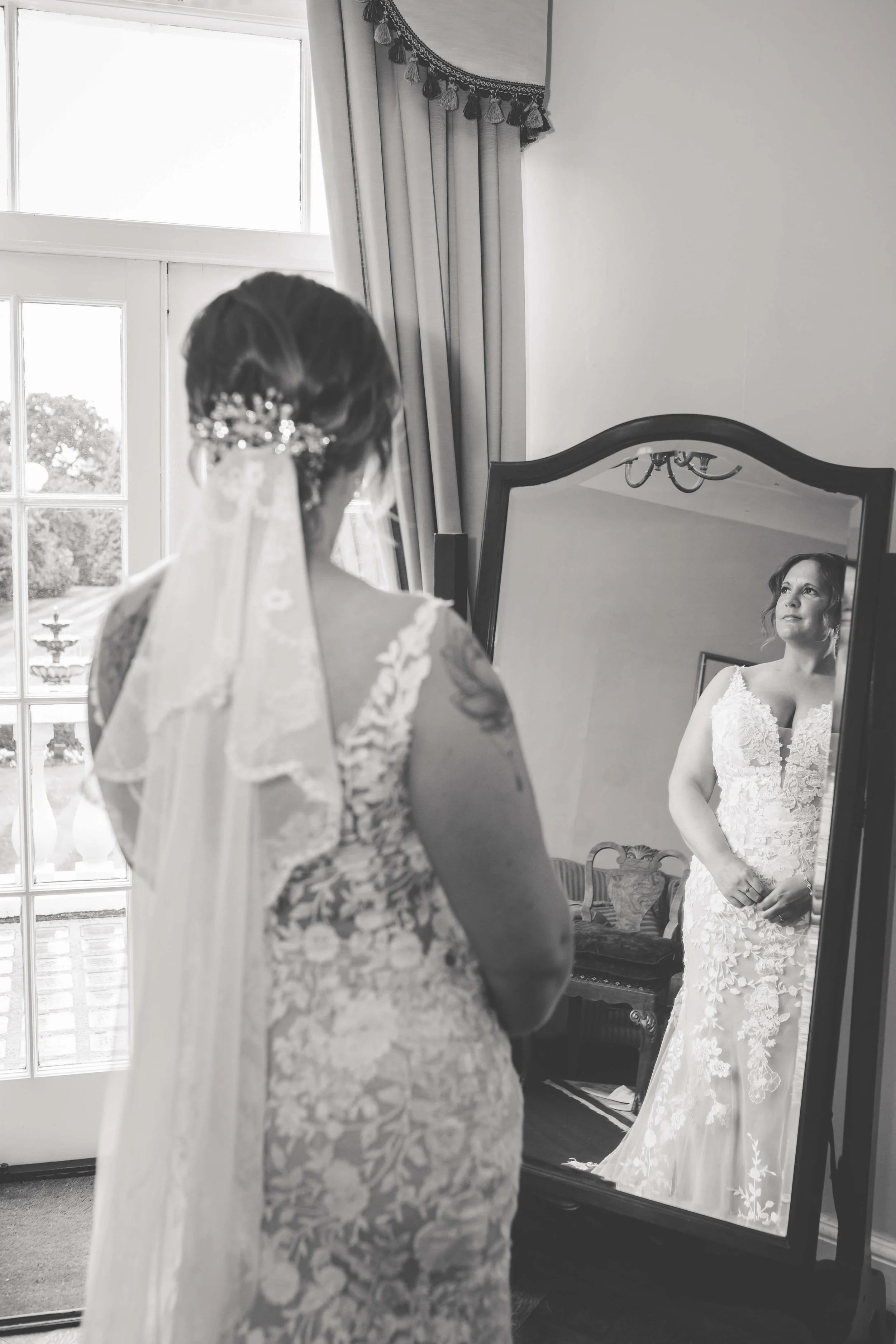 A bride in a lace wedding dress standing in front of a full-length mirror, looking at her reflection, with a window and curtains in the background.