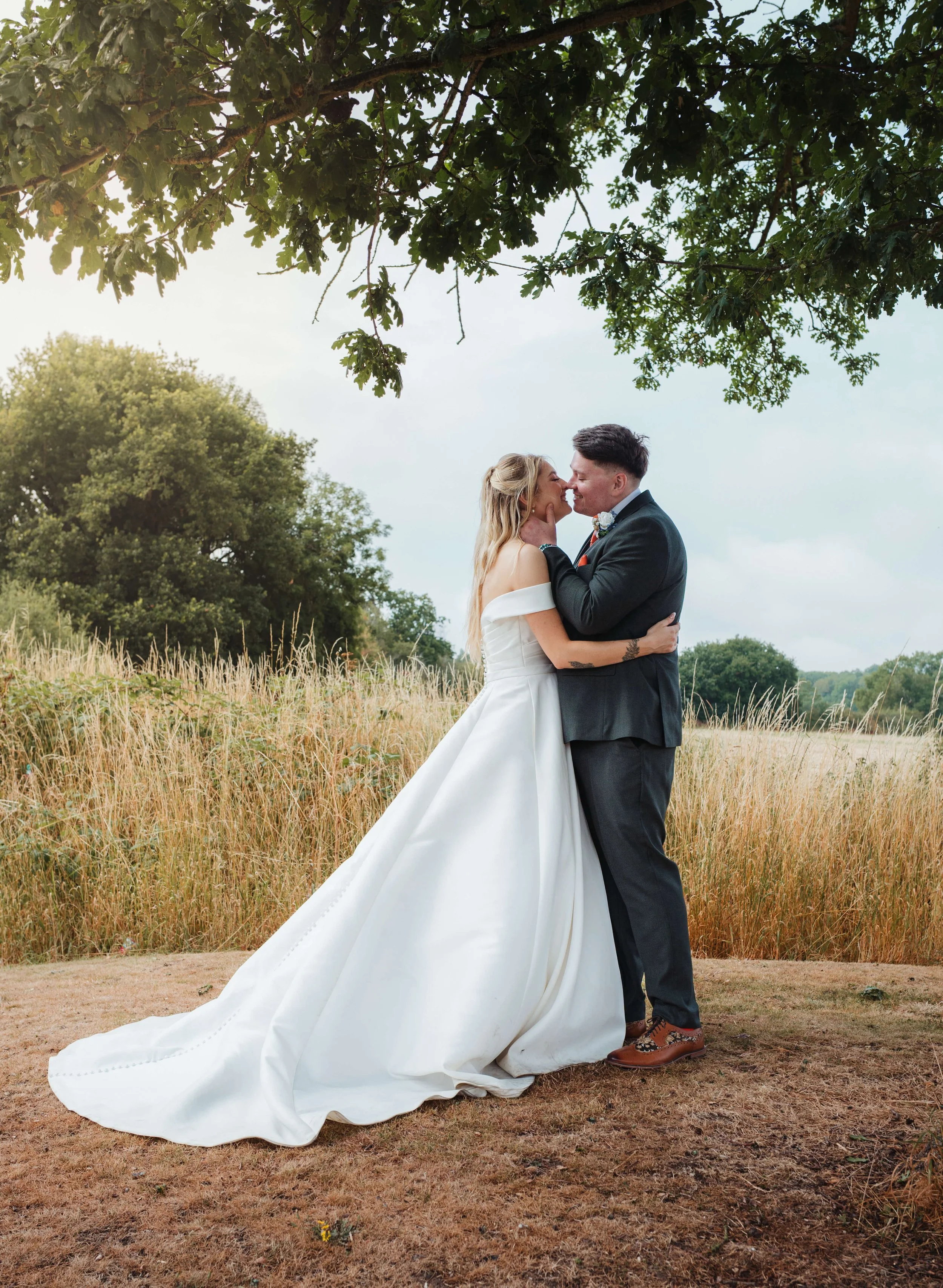 A bride and groom embrace outdoors in a field, under a large tree, with tall grass and green trees in the background, during their wedding.