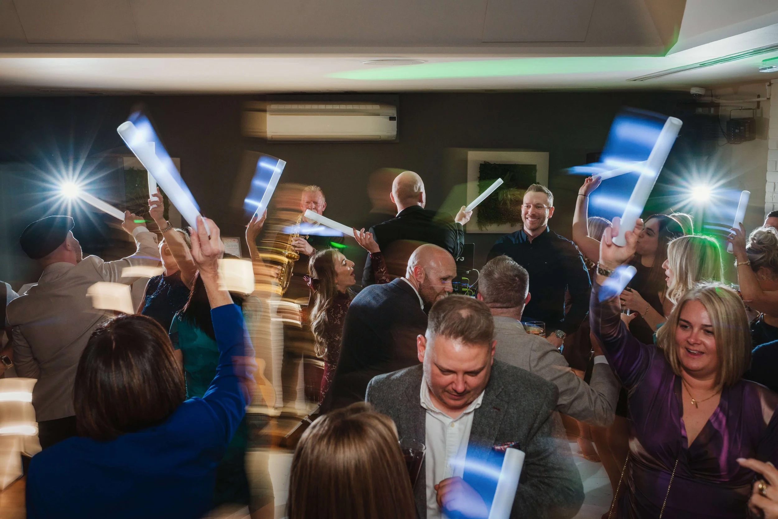 People dancing and celebrating at a party, with some holding glowing light sticks, in a lively indoor setting.