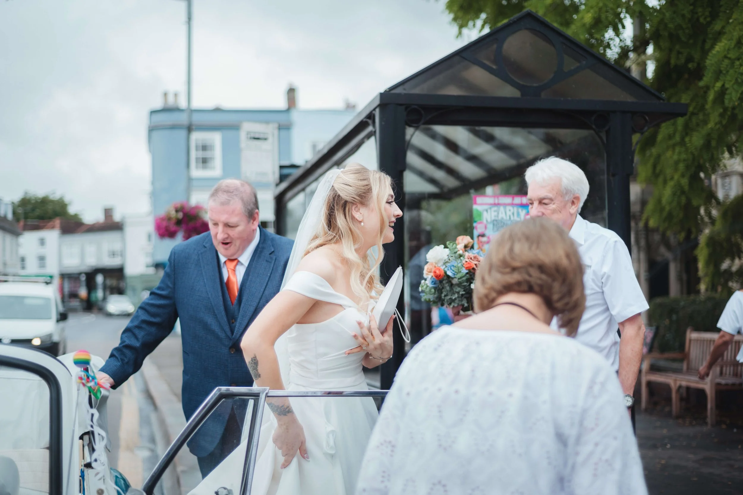A bride in a white wedding dress talking with an older man holding a bouquet of flowers at a bus stop, with a woman in white and a man in a suit nearby.