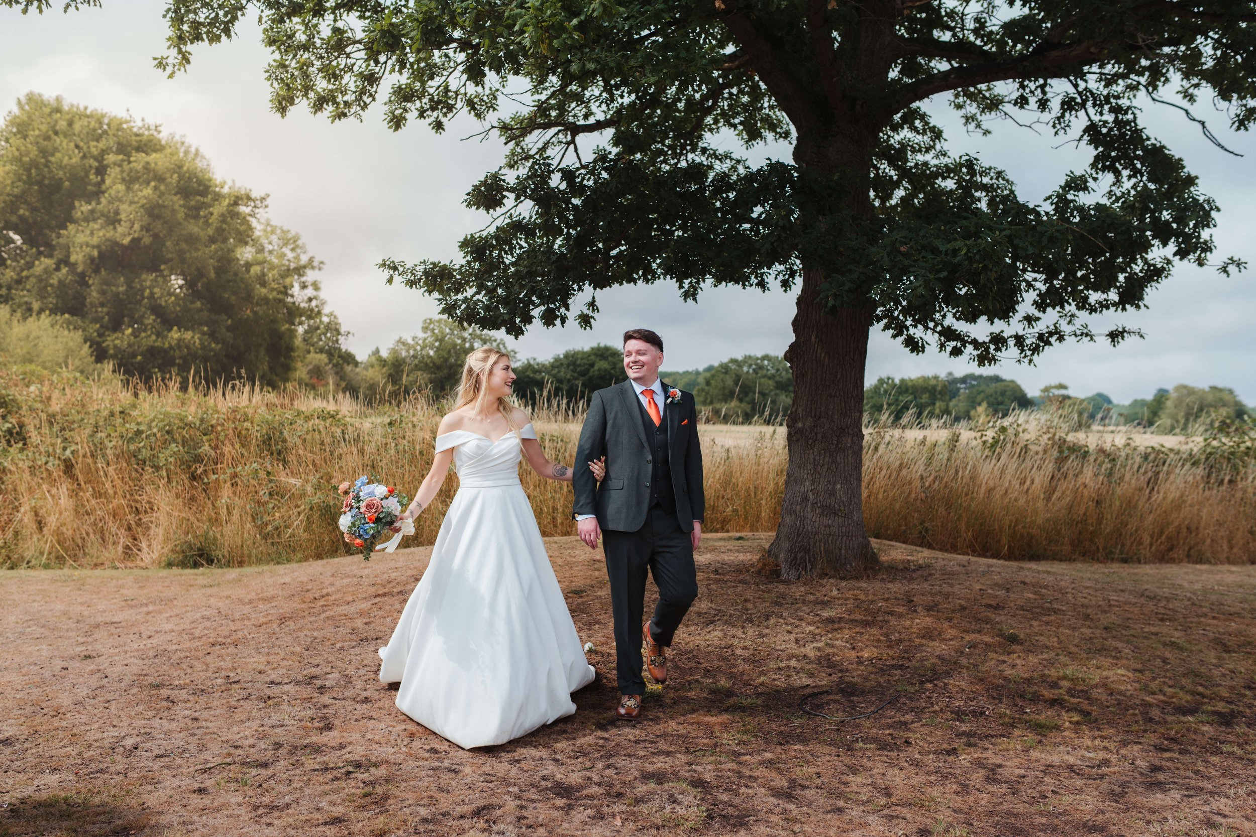 A bride and groom walking arm in arm outdoors near a large tree, smiling and looking at each other. The bride is wearing a white off-shoulder wedding gown, holding a bouquet of flowers. The groom is dressed in a dark suit with an orange tie and bouto