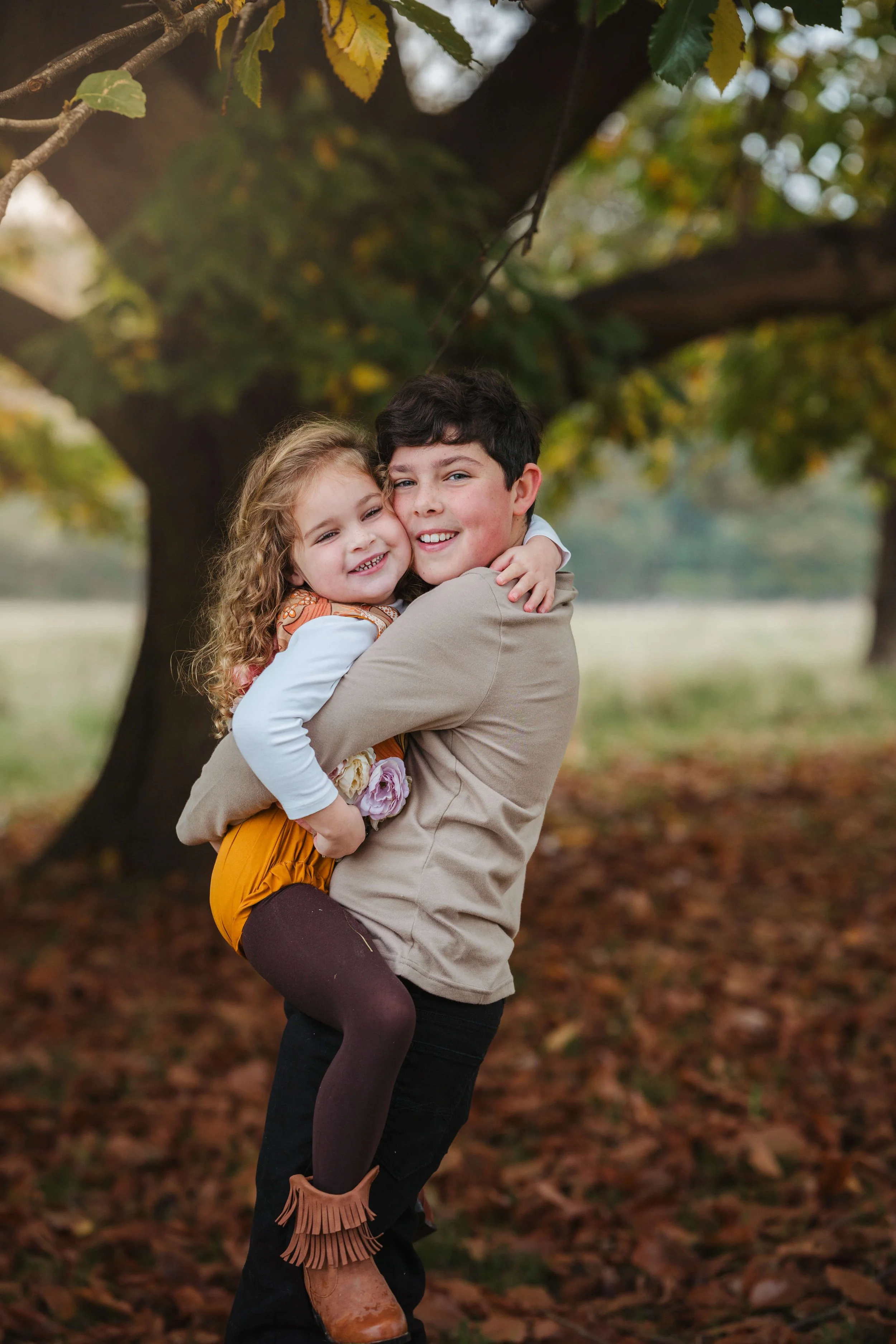 A young boy lifting a smiling girl in his arms outdoors during fall, with trees and fallen leaves in the background.