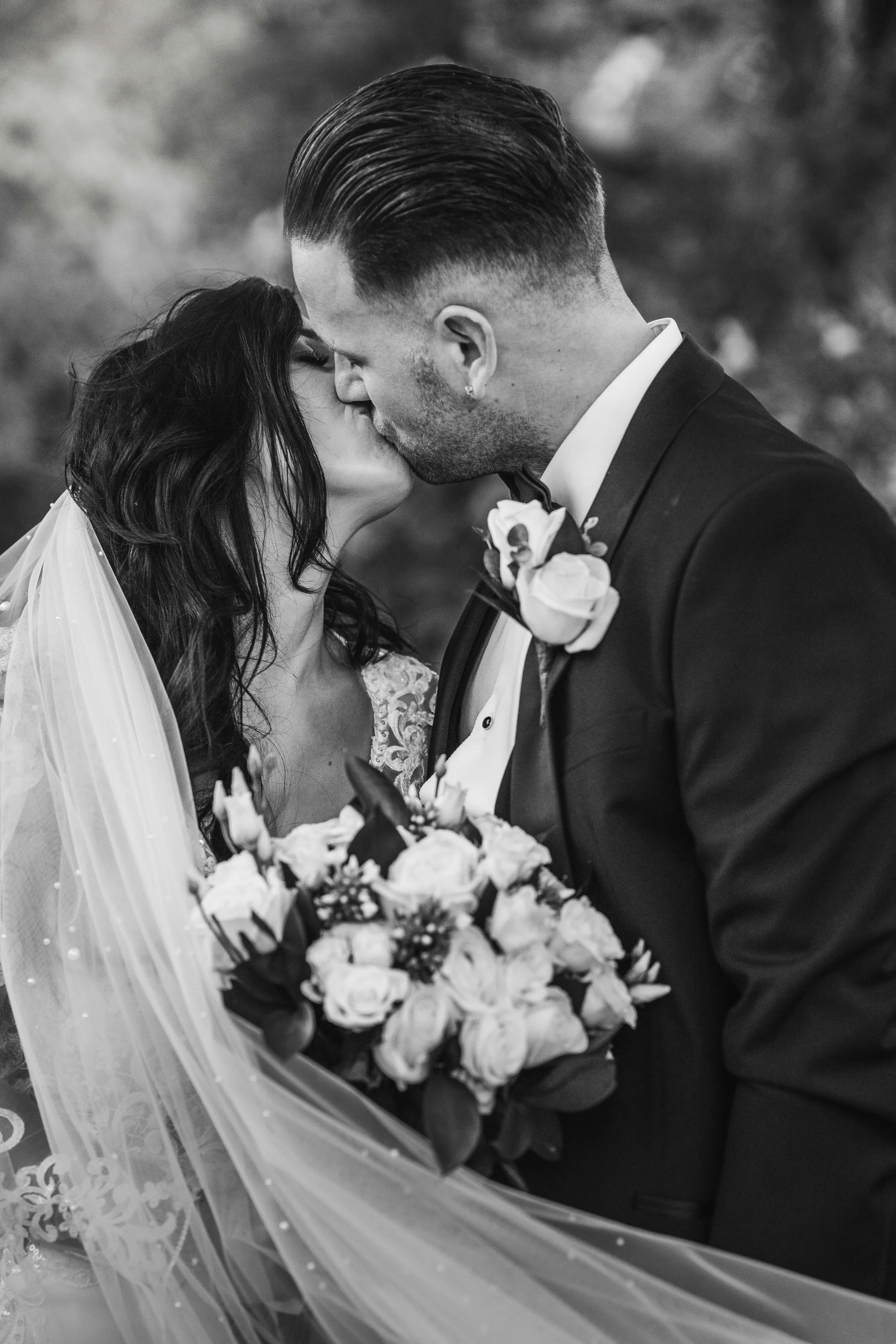 A black and white photo of a bride and groom sharing a kiss, with the bride holding a bouquet of flowers, both dressed in wedding attire.