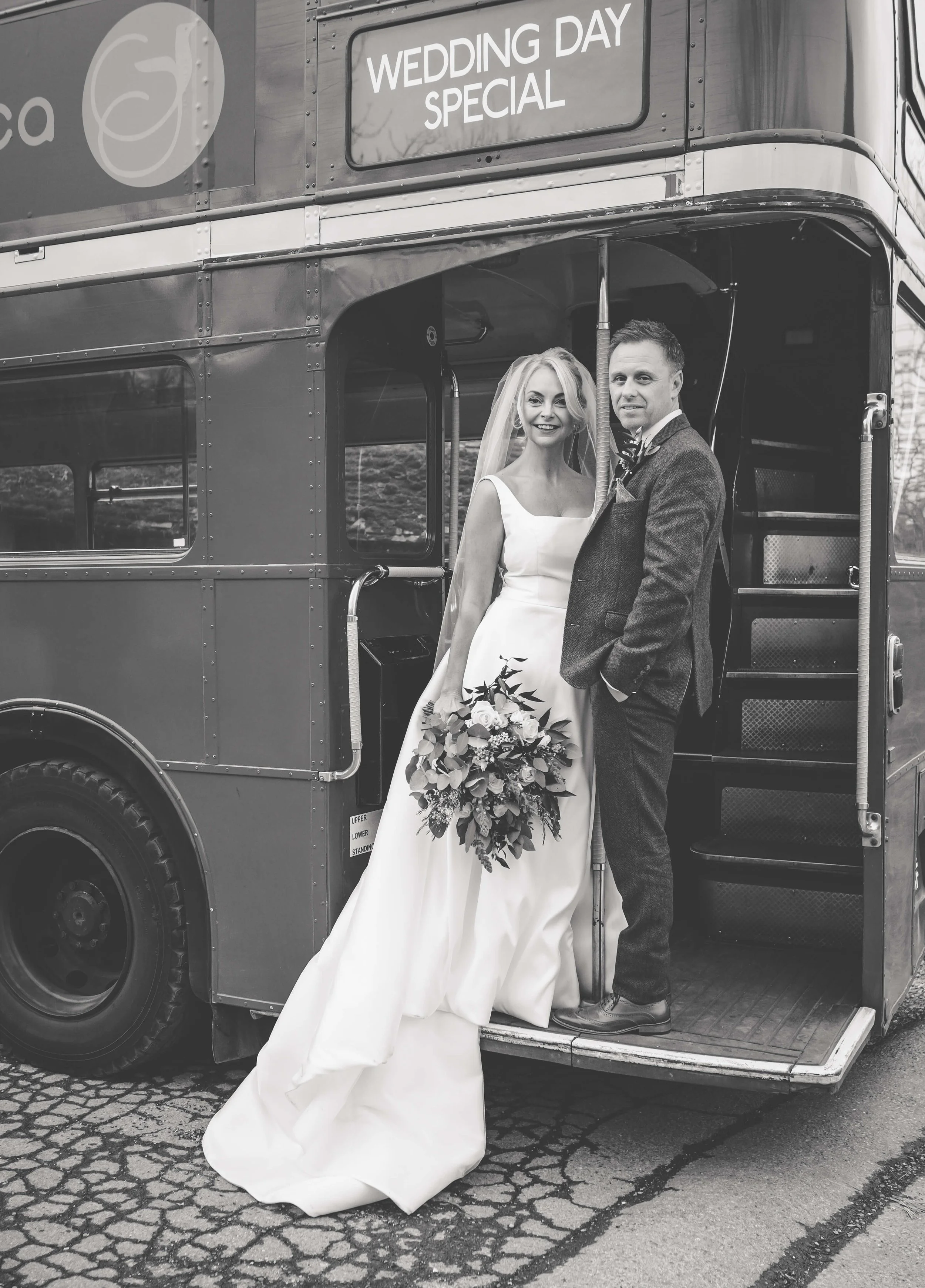 Black and white photo of a bride and groom standing in the doorway of a double-decker bus labeled "Wedding Day Special." The bride is holding a bouquet of flowers and wearing a long wedding dress with a veil. The groom is dressed in a suit with a bow
