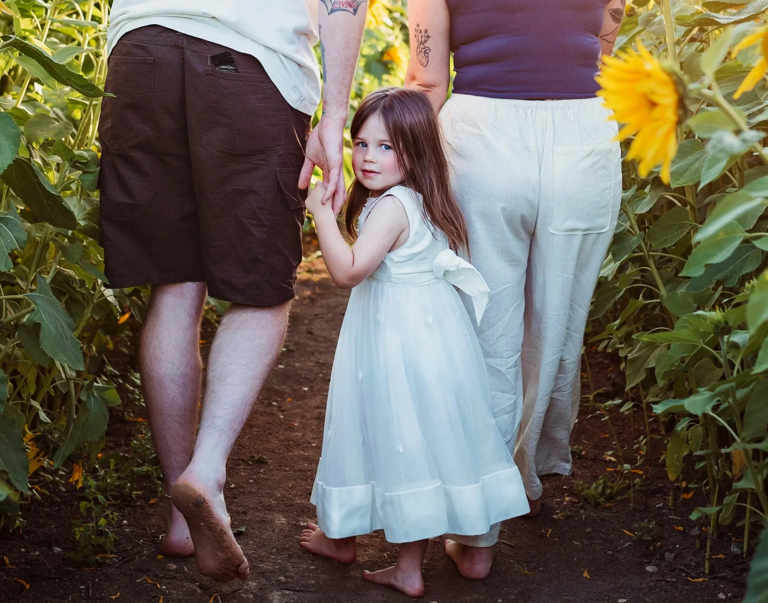 A young girl in a white dress holding hands with two adults, walking barefoot through a sunflower field.