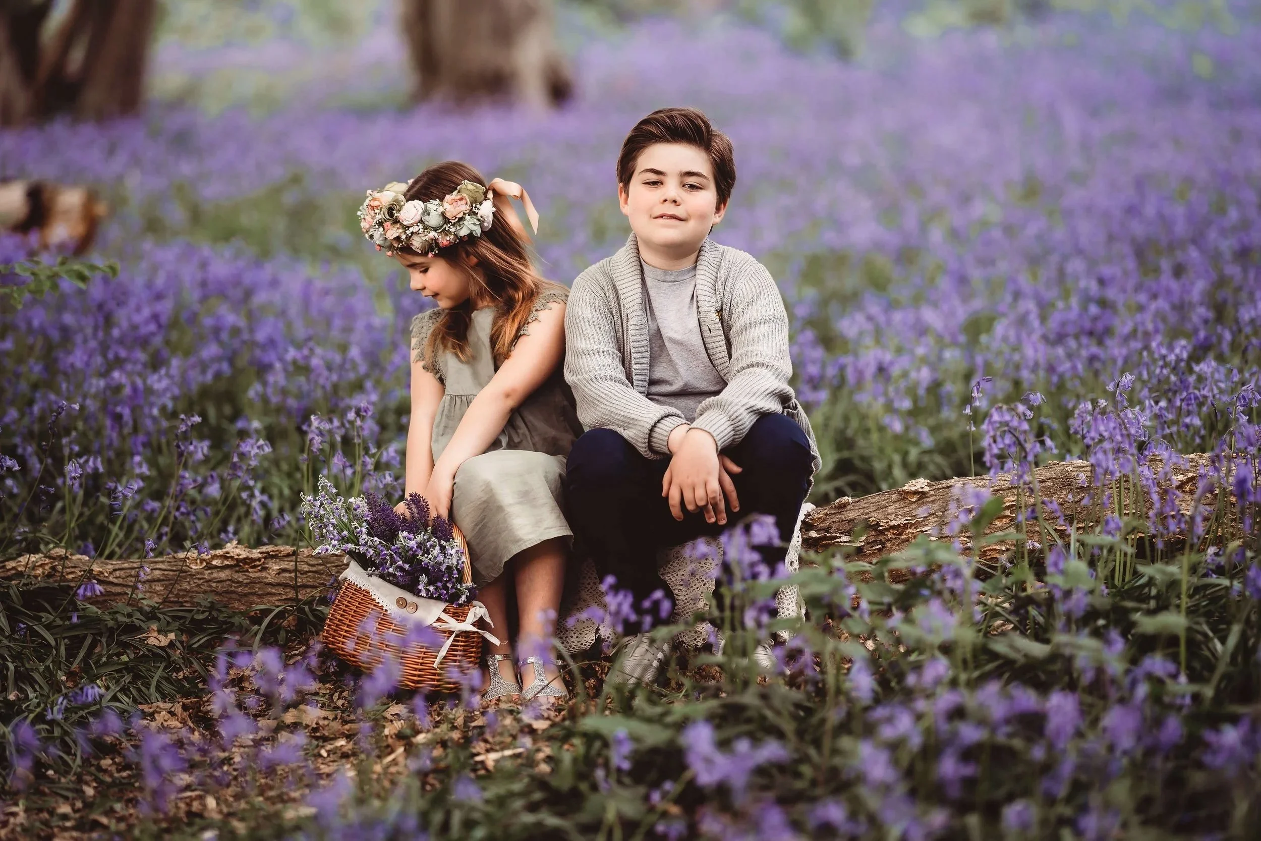 A boy and a girl sitting on a log in a purple flower field, with the girl wearing a flower crown and holding a basket of flowers, and the boy with a grey sweater and dark pants.