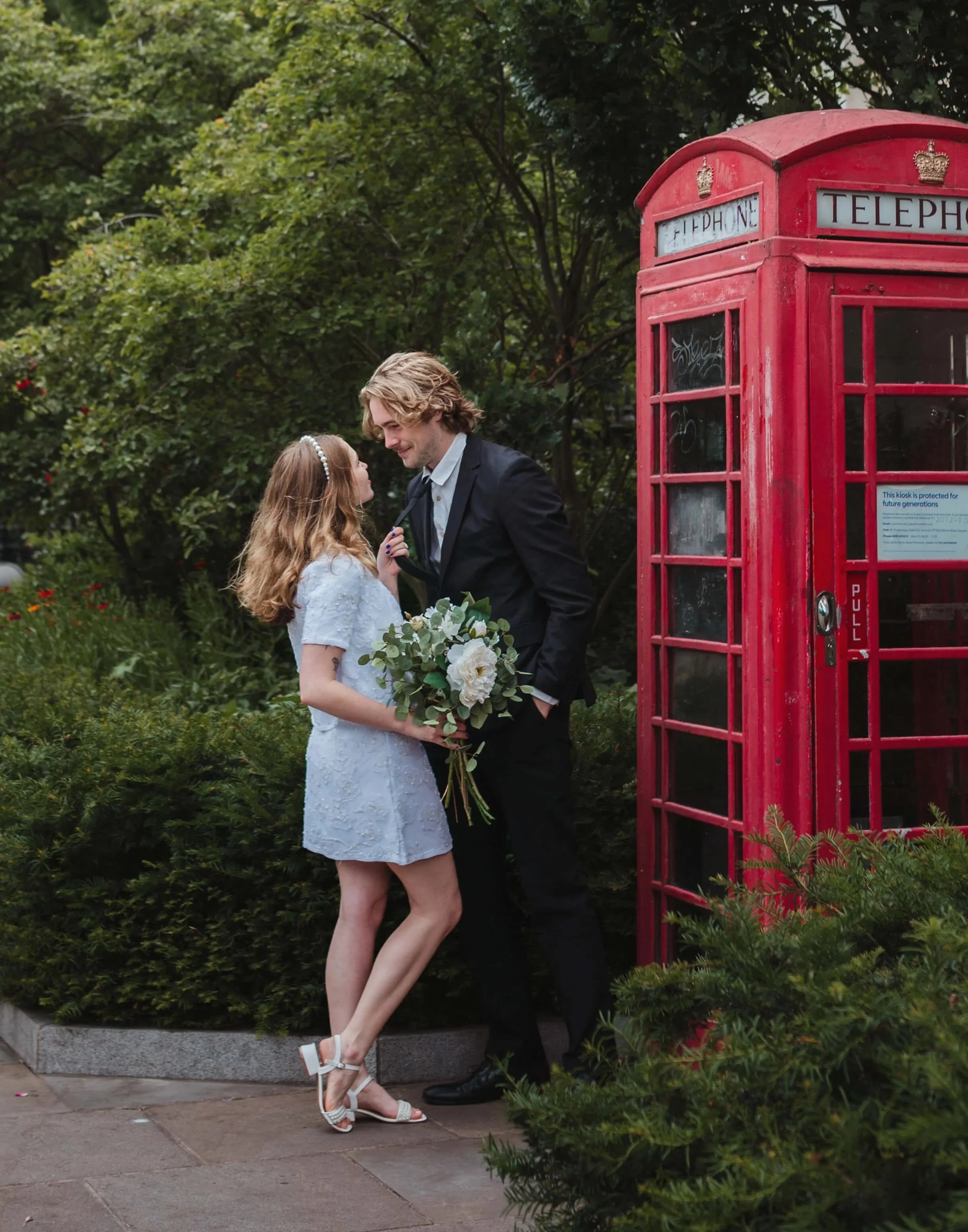 A couple dressed formally, the woman holding a bouquet, standing near a red British telephone booth in a green park.