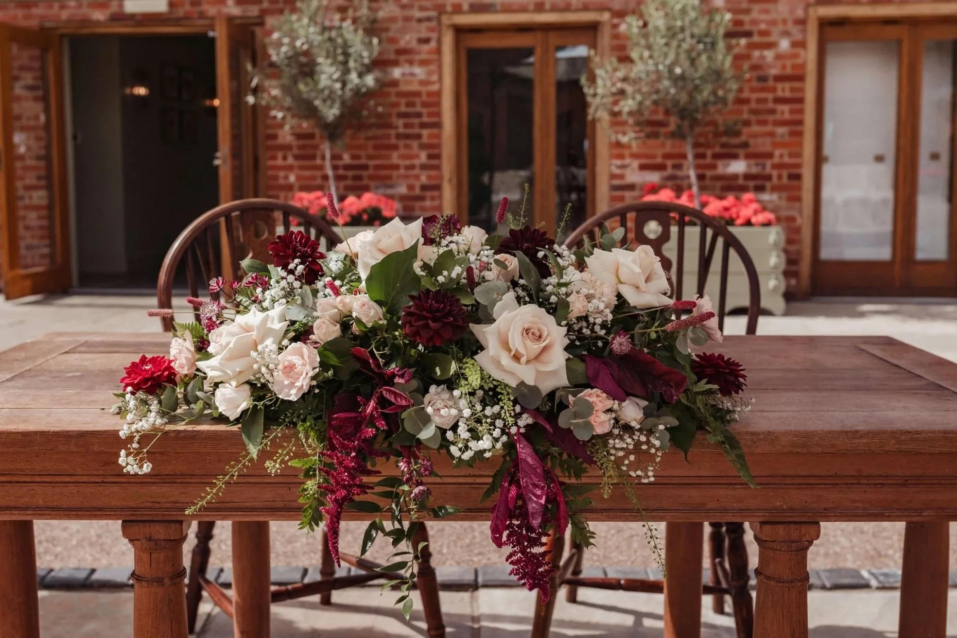 A floral centerpiece with white roses, burgundy flowers, and greenery on a wooden table outside a brick building with glass doors.