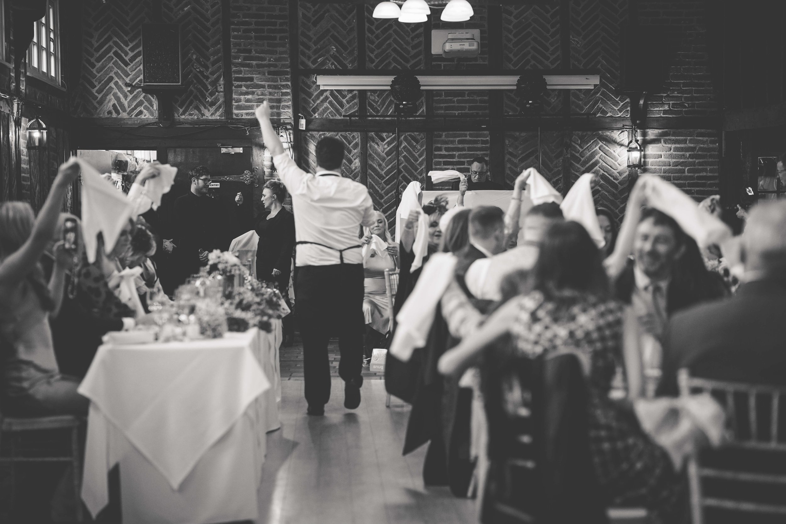 People sitting at tables with white tablecloths, attending a celebration or dinner event in a brick-walled venue. A waiter in white shirt and black apron walks through the hall, while other people wave white napkins or cheer.