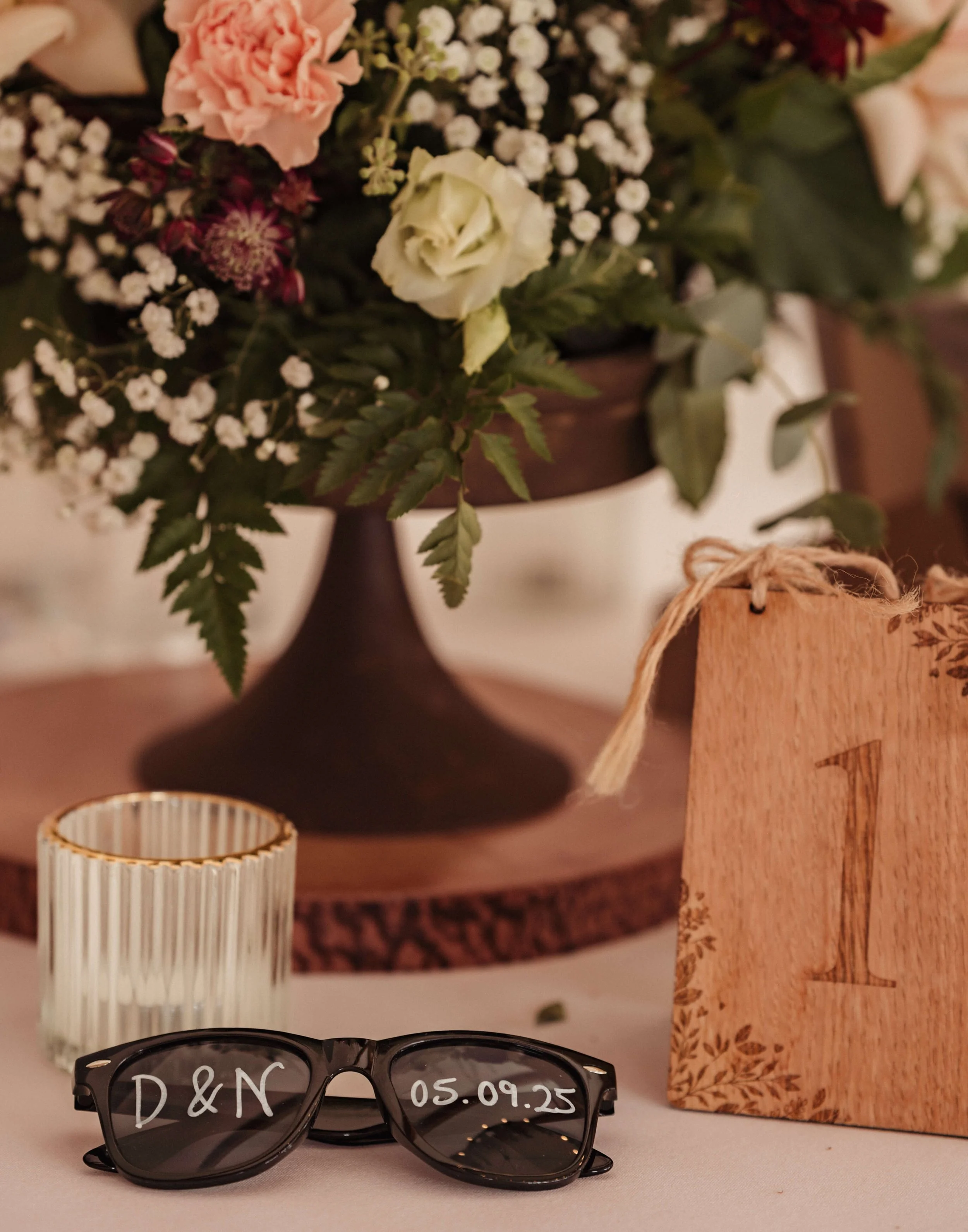 A floral arrangement with pink, white, and burgundy flowers and greenery in a dark wood pedestal vase, a small wooden table number sign with the number one and a floral border, a pair of black sunglasses with 'D & N' and the date '05.09.25' written o