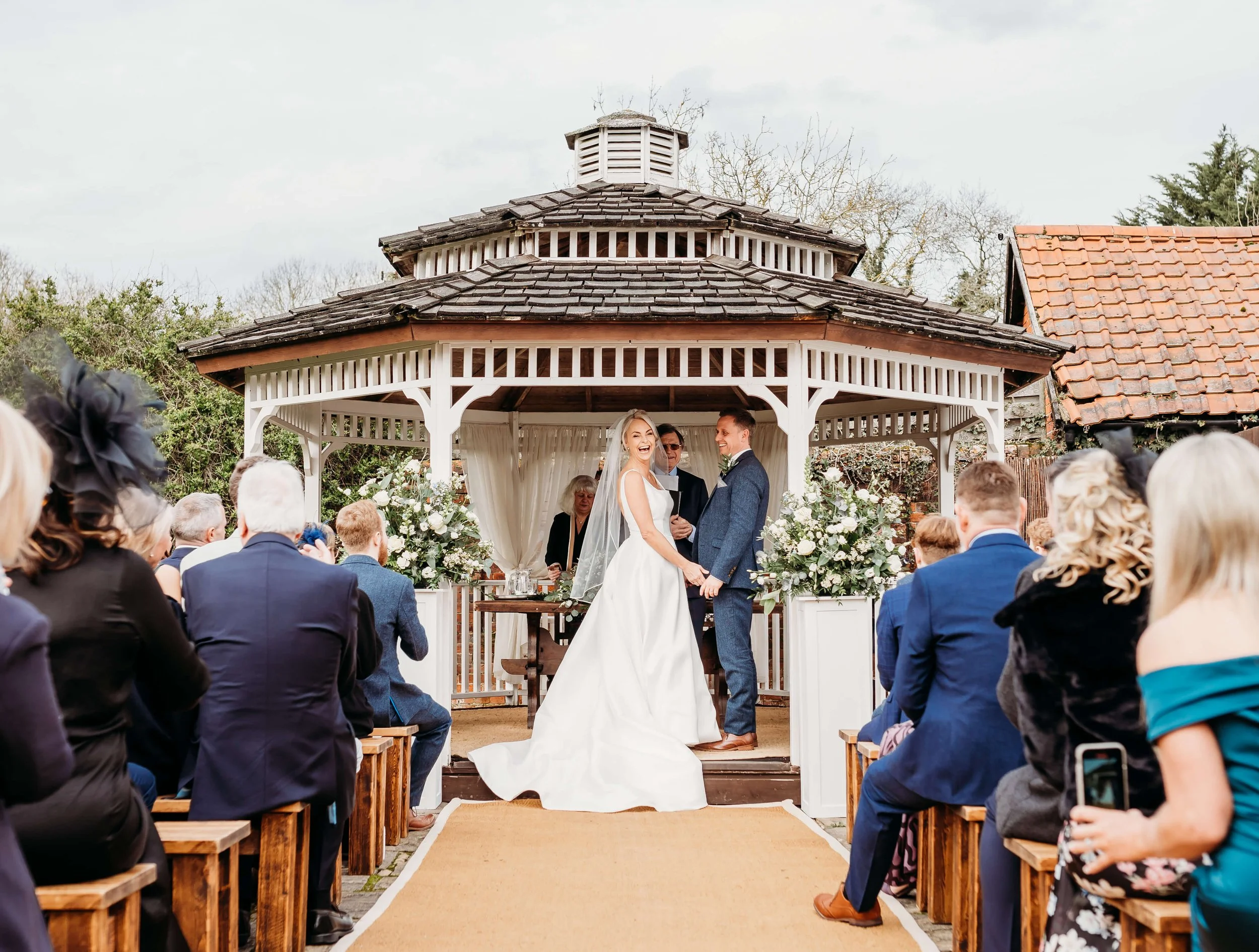 A wedding ceremony taking place in an outdoor gazeboo, with a bride and groom holding hands and smiling at each other, surrounded by guests seated on wooden benches, with floral arrangements and an officiant present.