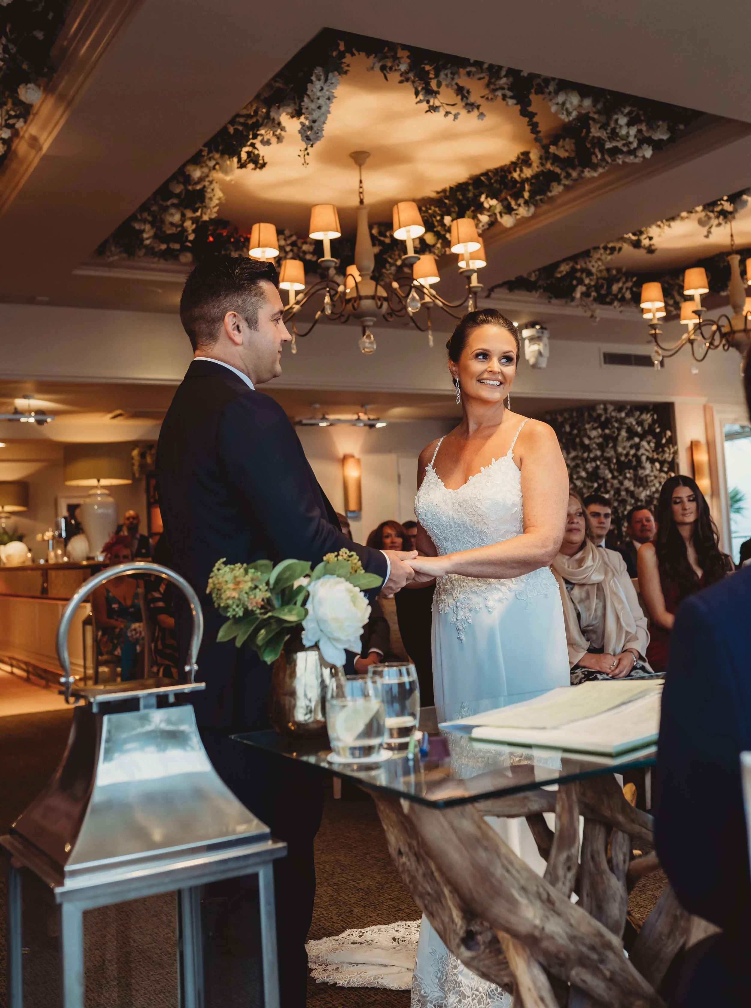 A bride and groom holding hands during a wedding ceremony indoors, with guests seated in the background and floral decorations overhead.