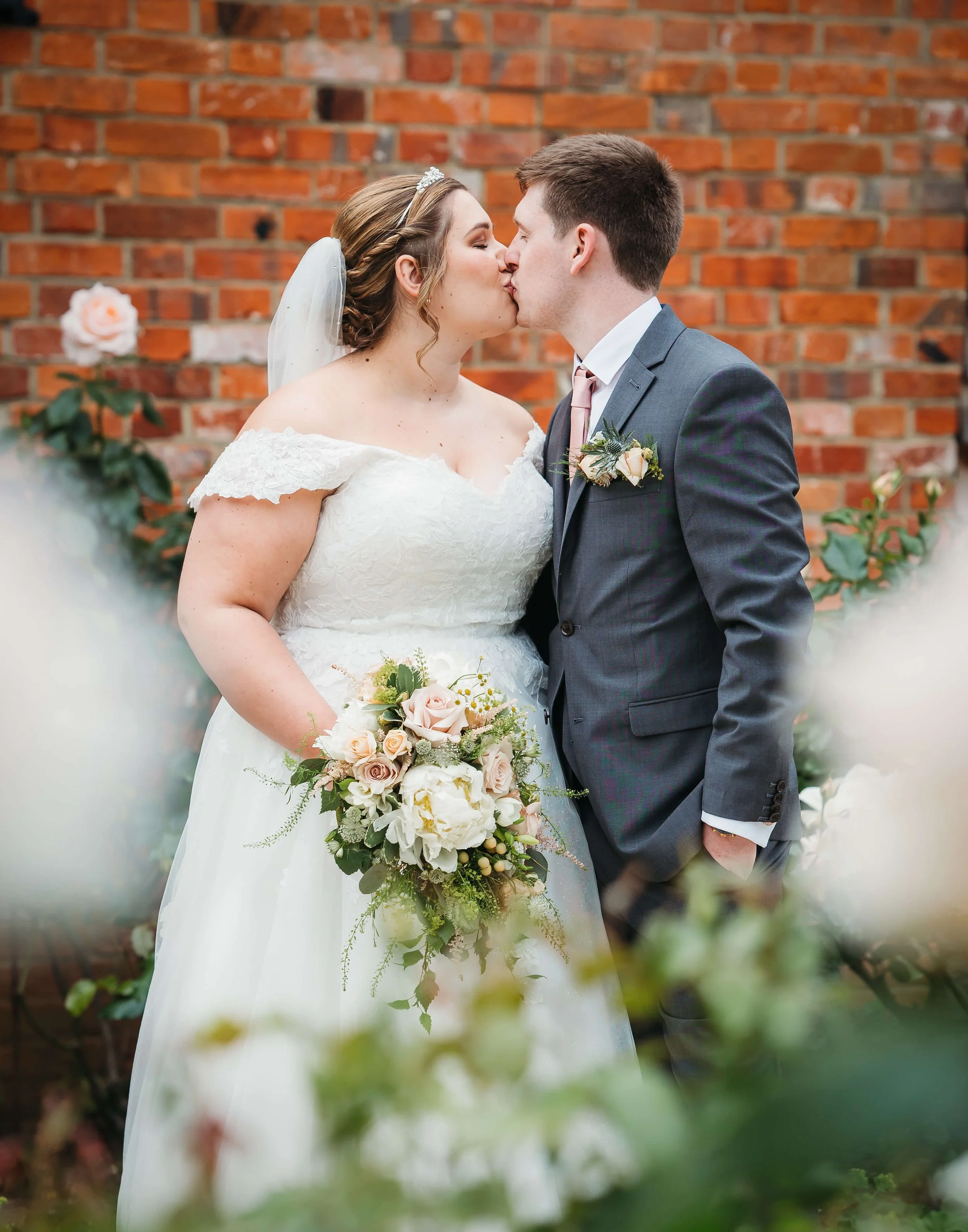 A bride and groom sharing a kiss at their wedding, with a brick wall and floral decorations in the background. The bride is holding a bouquet of light pink and white flowers, wearing a white lace off-shoulder wedding dress and veil. The groom is dres