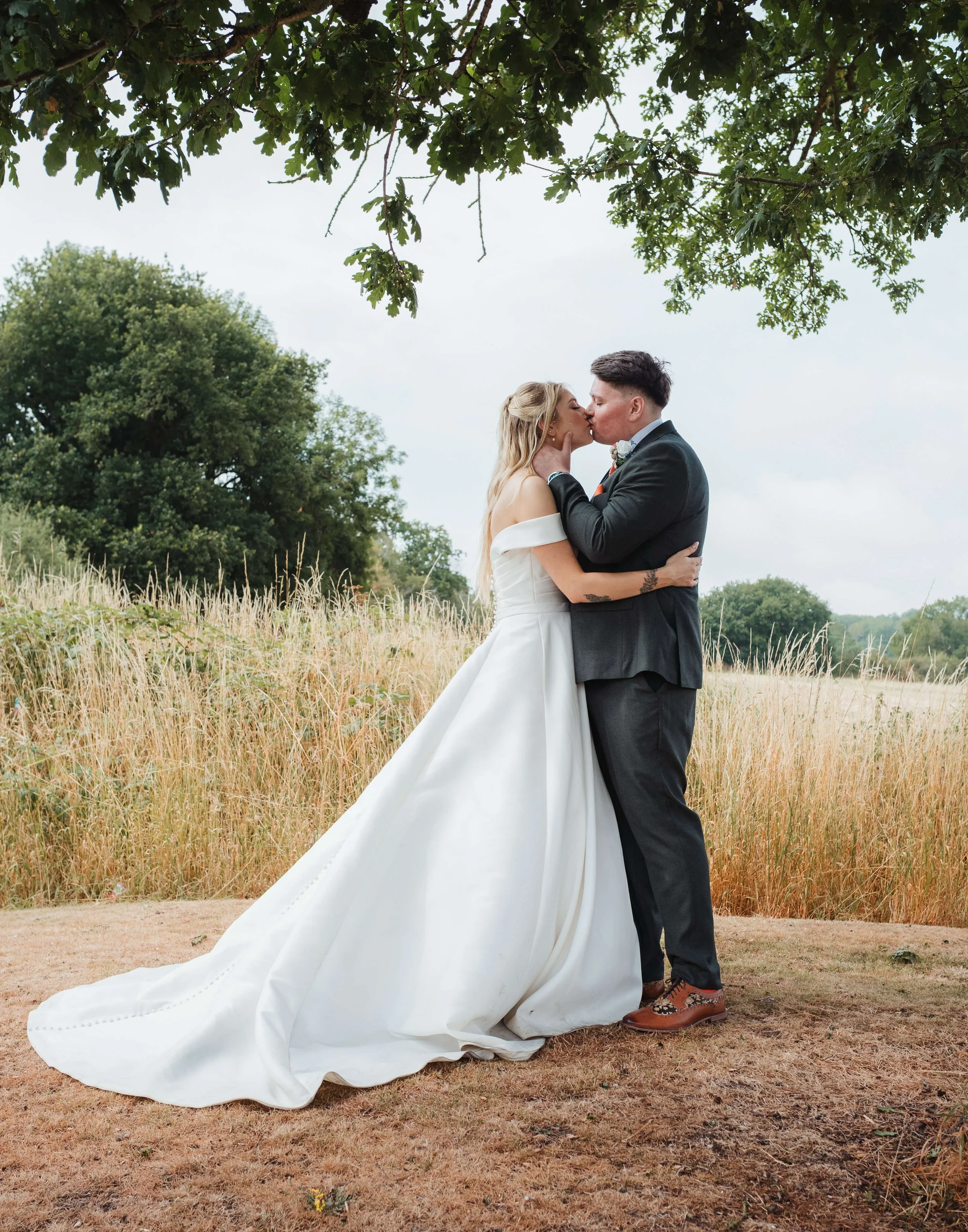 A couple in wedding attire sharing a kiss outdoors in a field with trees in the background.