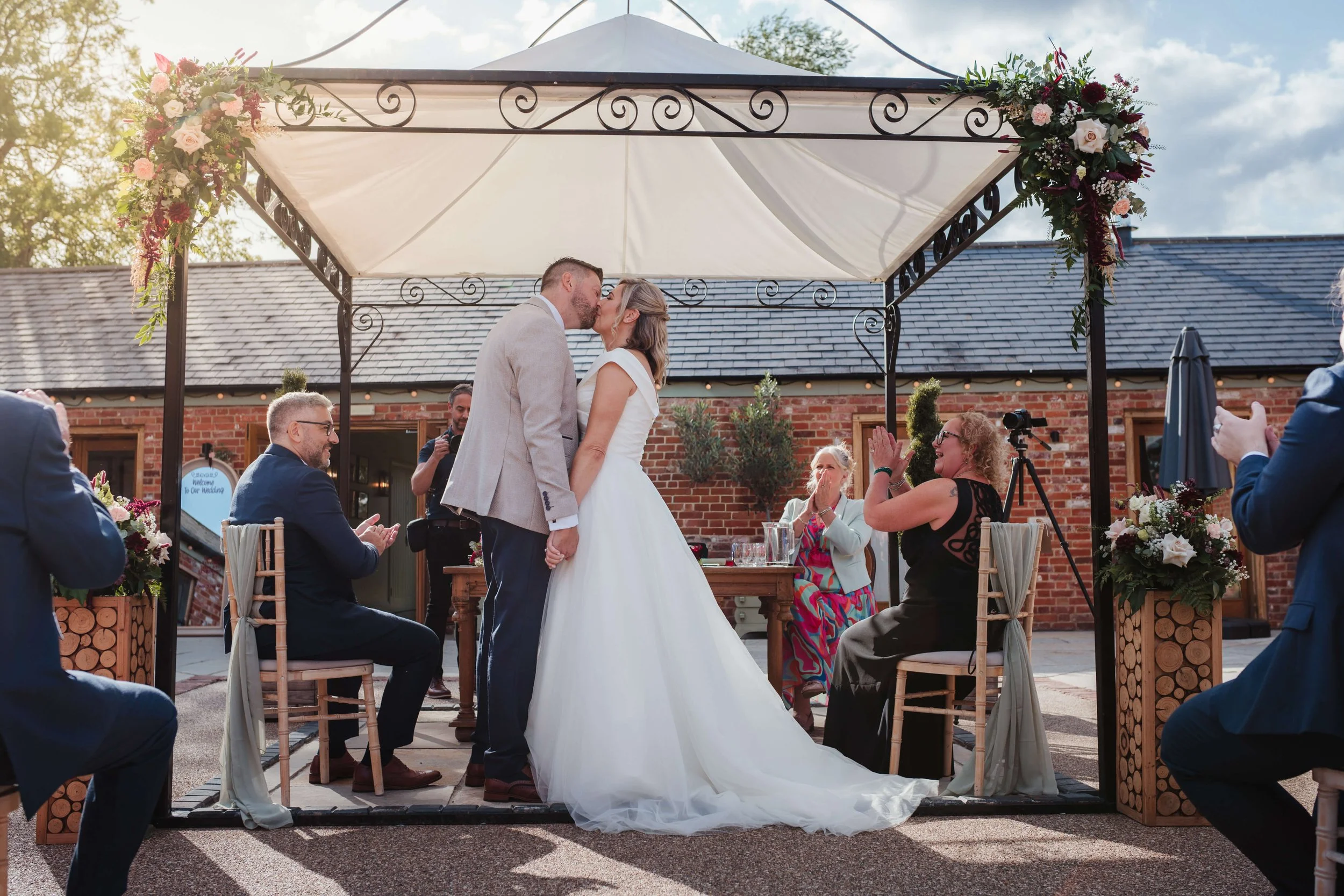 A couple kissing during their wedding ceremony under a decorated canopy, with guests and officiant present, outdoors during daytime.