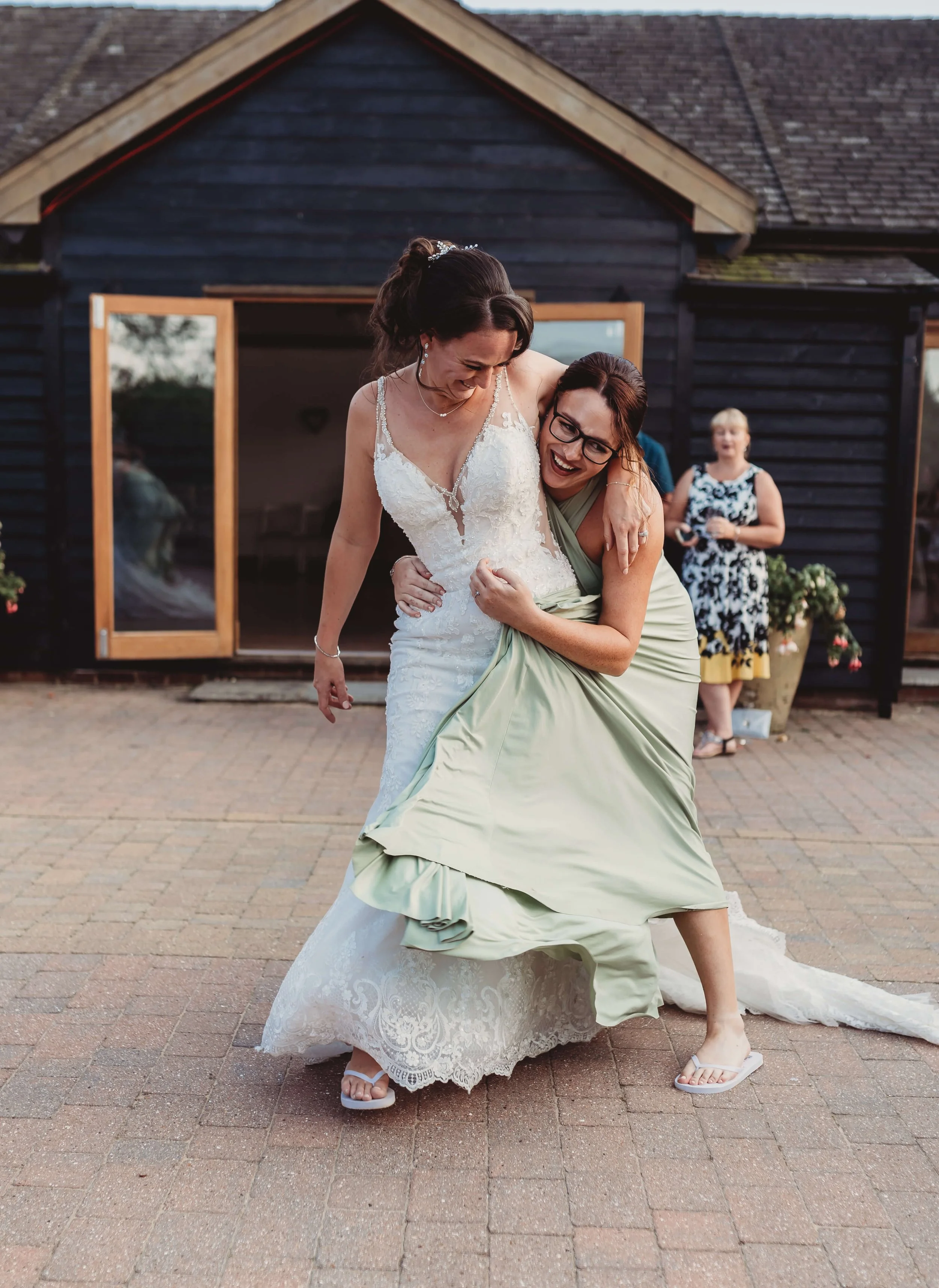 Two women, one in a wedding dress and the other in a light green dress, are dancing and laughing outside a house. A third person is in the background.