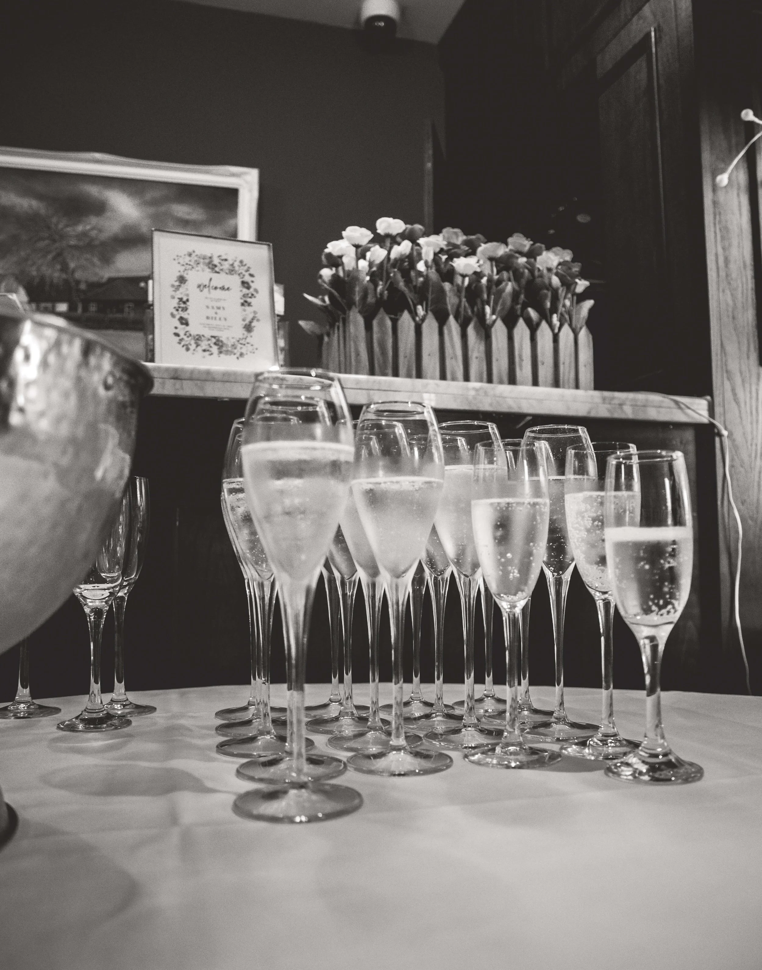 Multiple champagne glasses filled with sparkling wine on a table, with a flower arrangement and framed picture in the background.