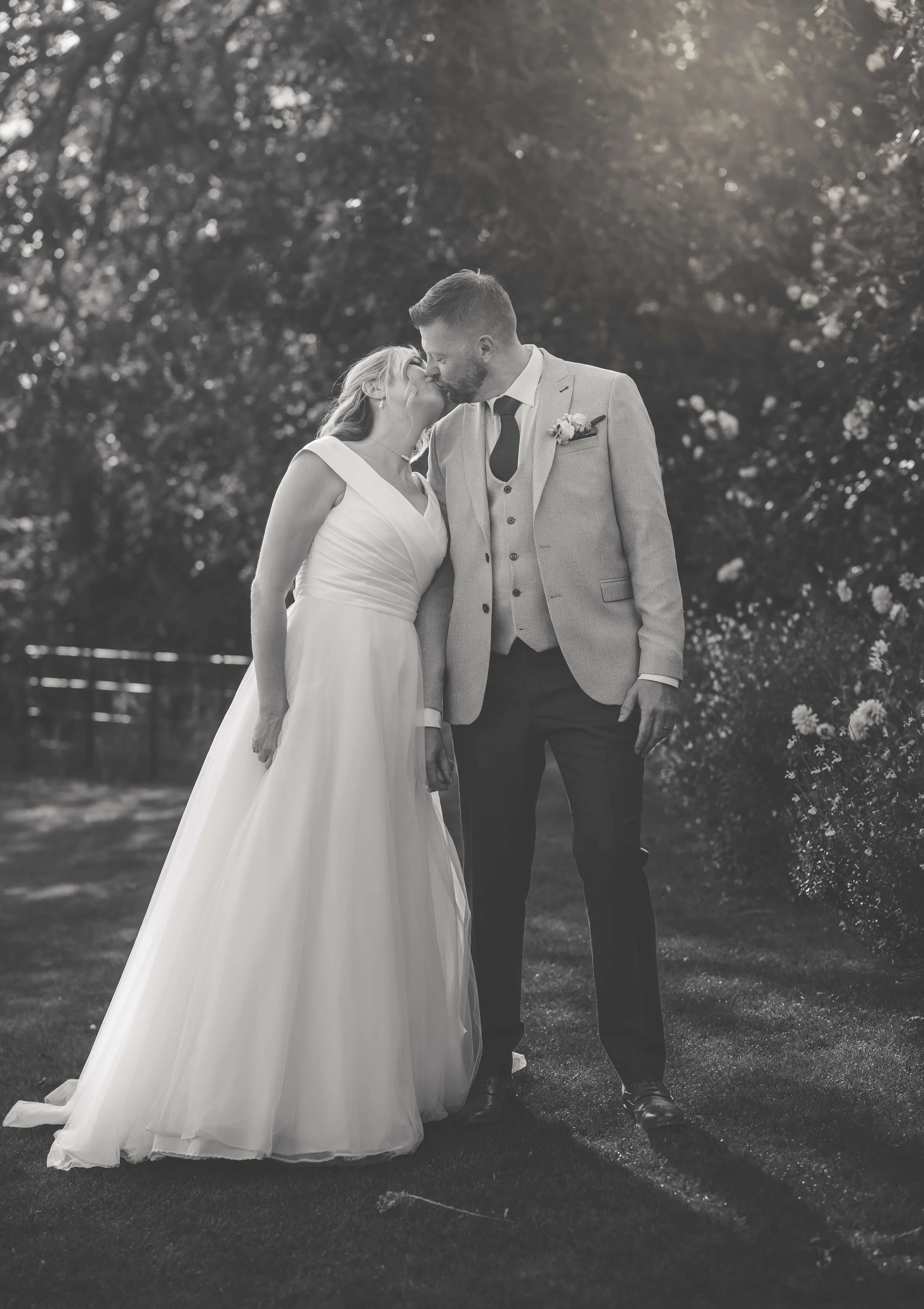 A bride and groom sharing a kiss outdoors during their wedding, dressed in formal wedding attire, with trees in the background.