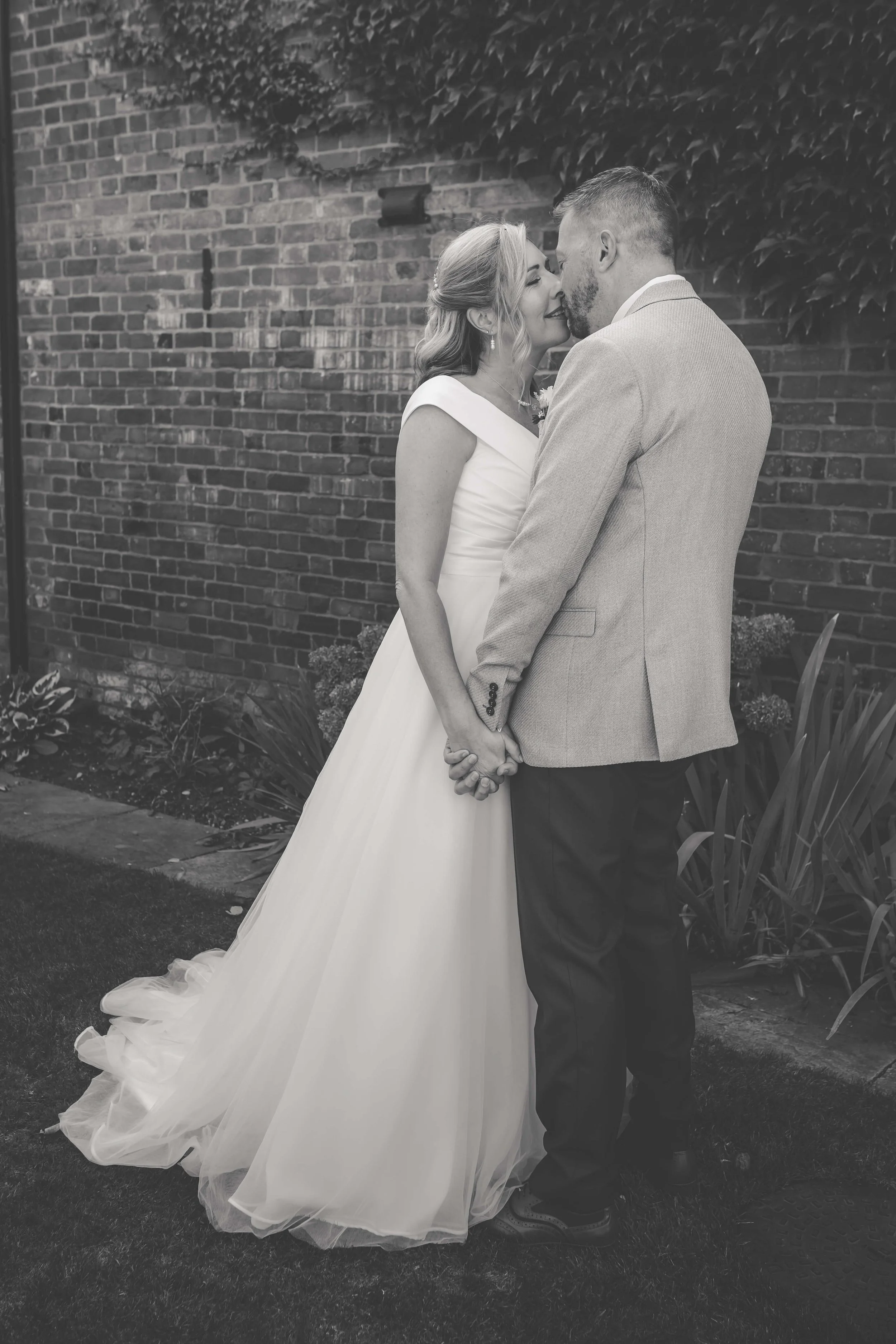 A bride and groom standing close together, holding hands, outdoors against a brick wall with foliage, about to kiss or touching noses.