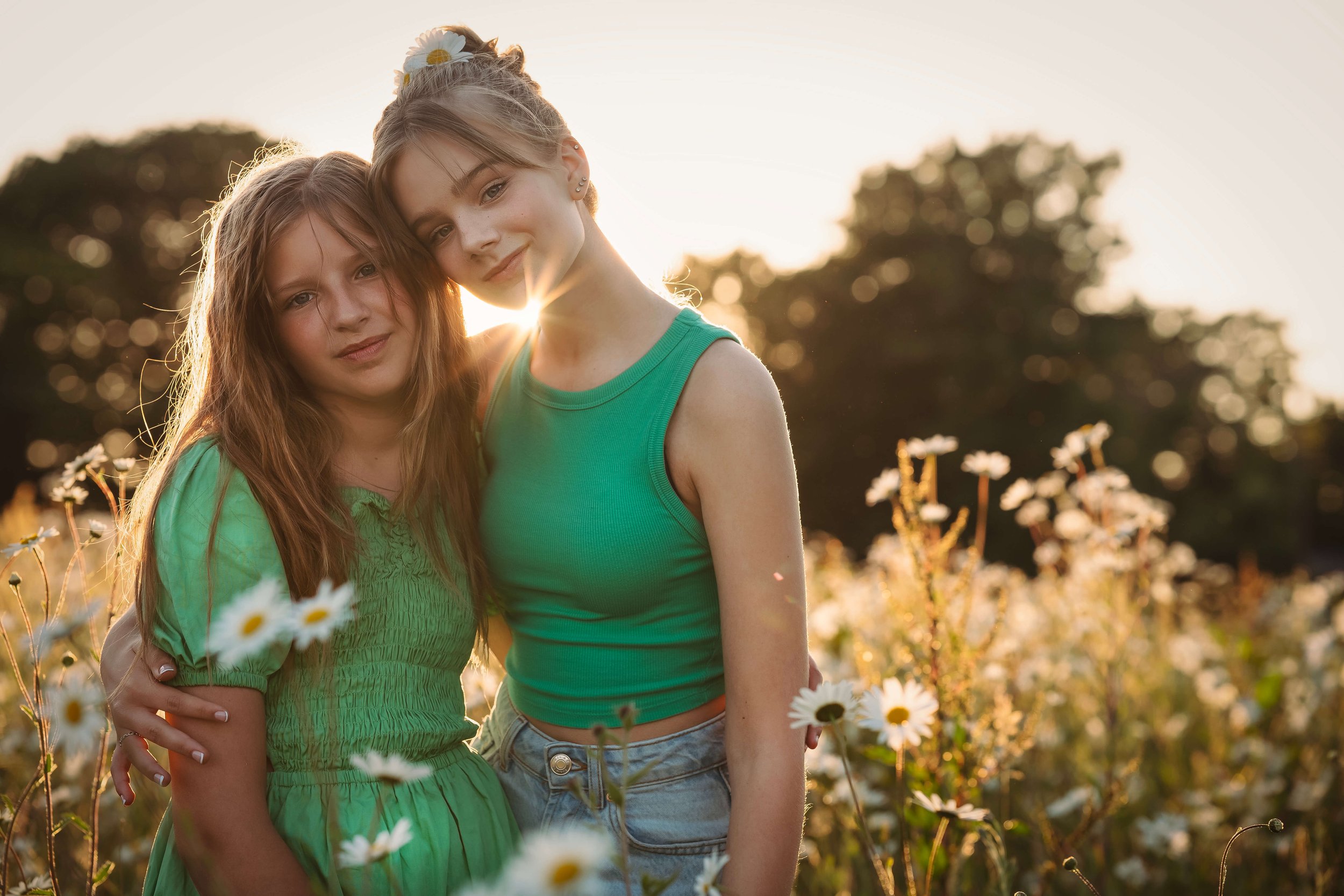 Two young girls standing close in a field of daisies during sunset, with one girl wearing a green dress and the other in a green sleeveless top.