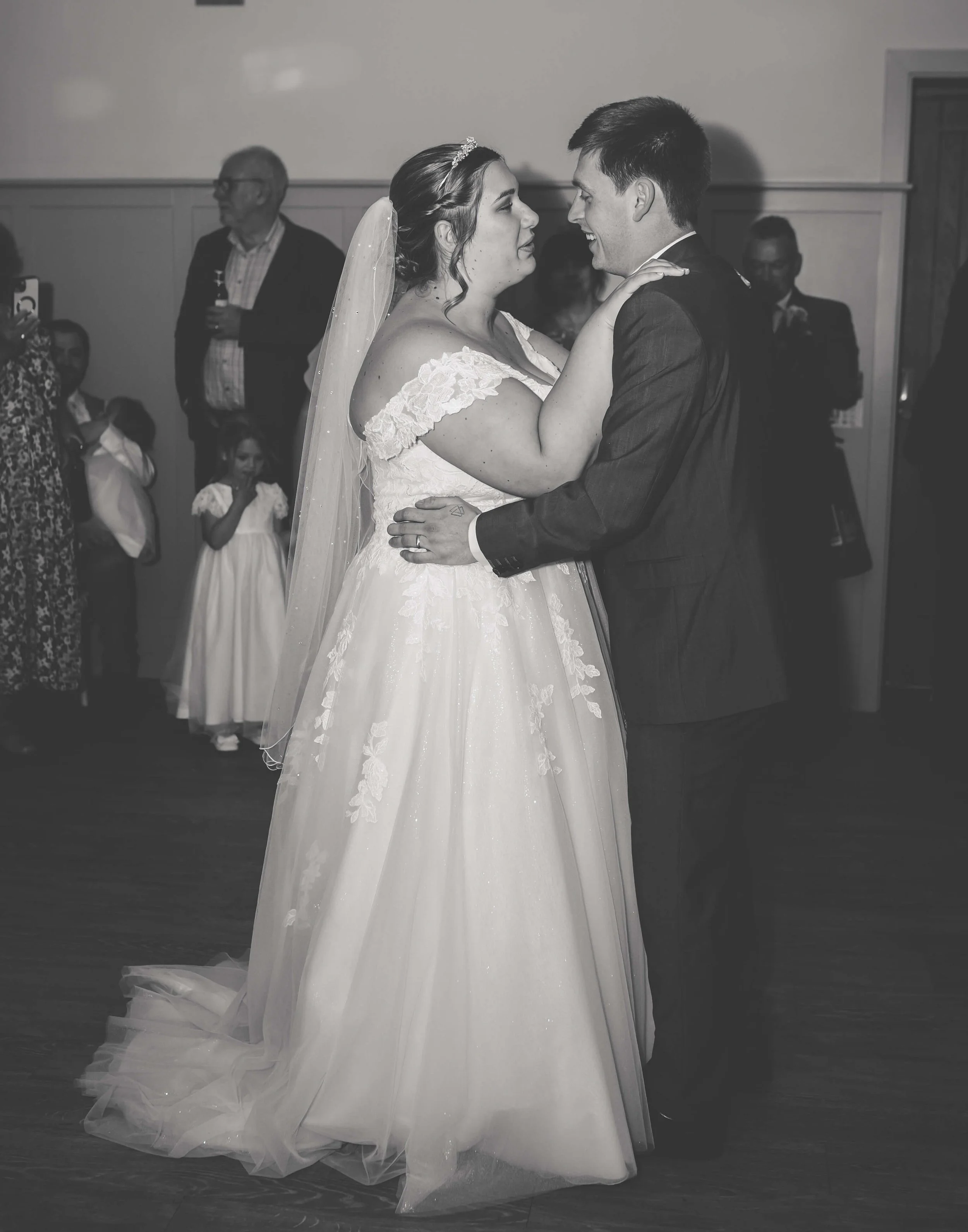 A black-and-white photo of a bride and groom dancing at their wedding reception, with guests watching.