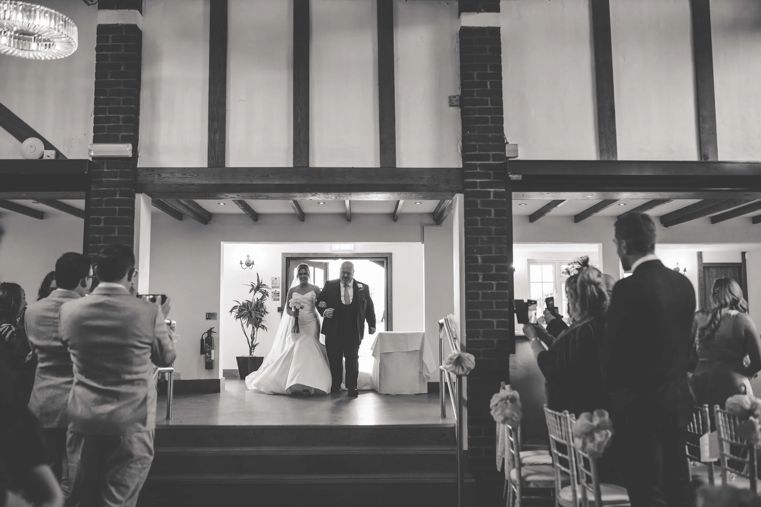 A bride walking down the aisle with an older man, likely her father, during a wedding ceremony in a rustic indoor venue with exposed brick and wooden beams, as guests take photos and watch.