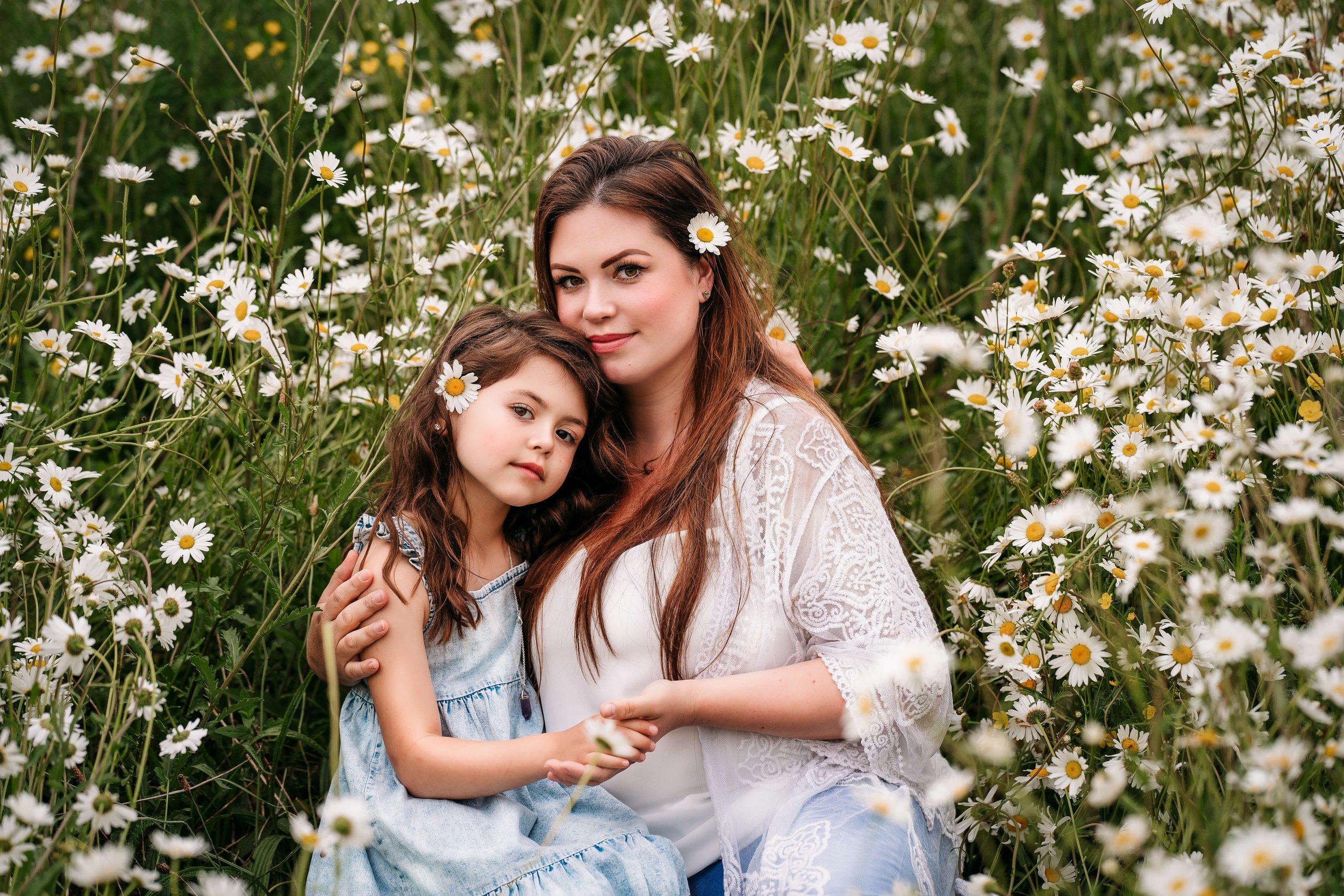 A woman and a girl sitting together in a field of white daisies, with daisies in their hair, looking at the camera.
