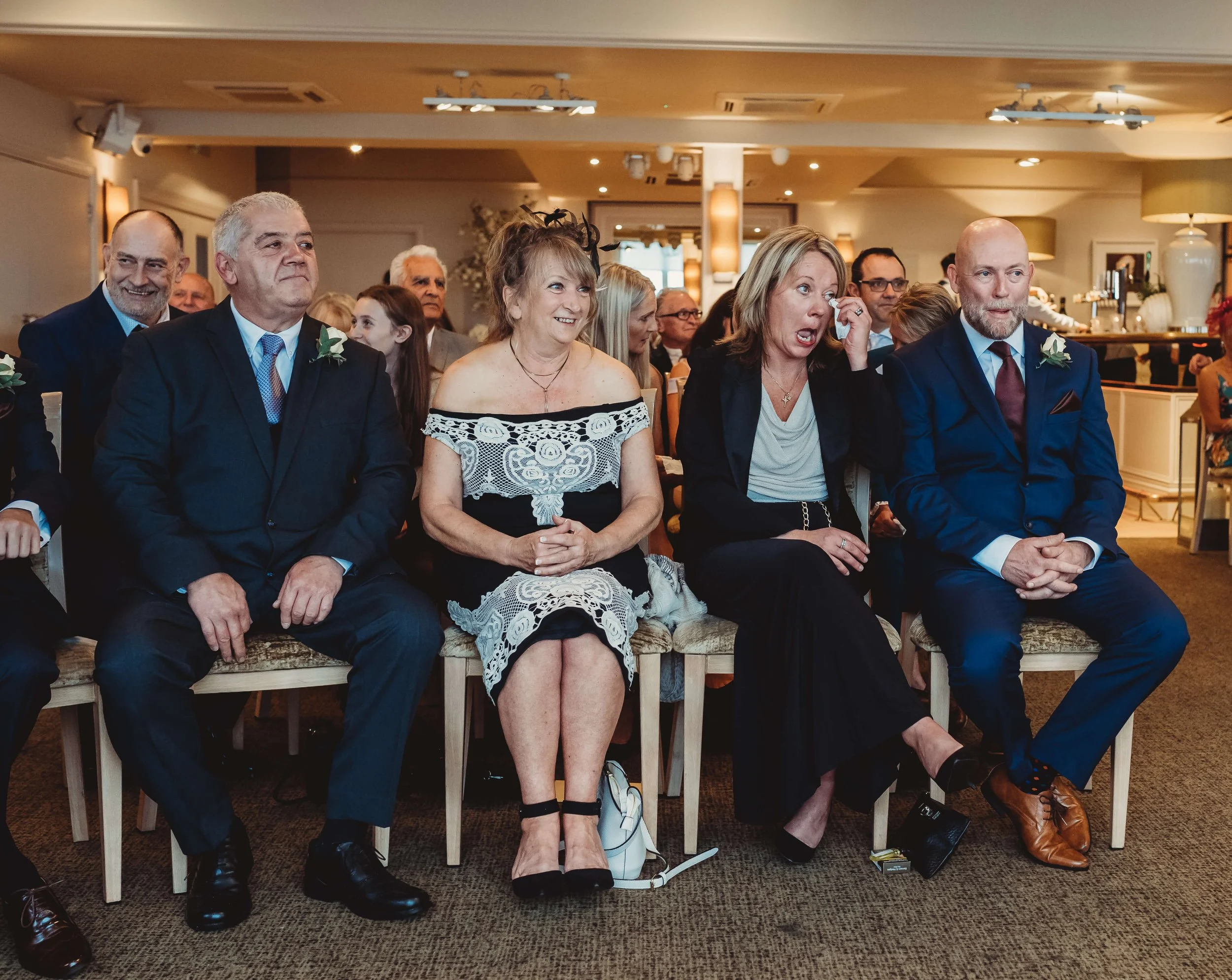 Group of people dressed in formal attire at a wedding or formal event, seated in a banquet hall.