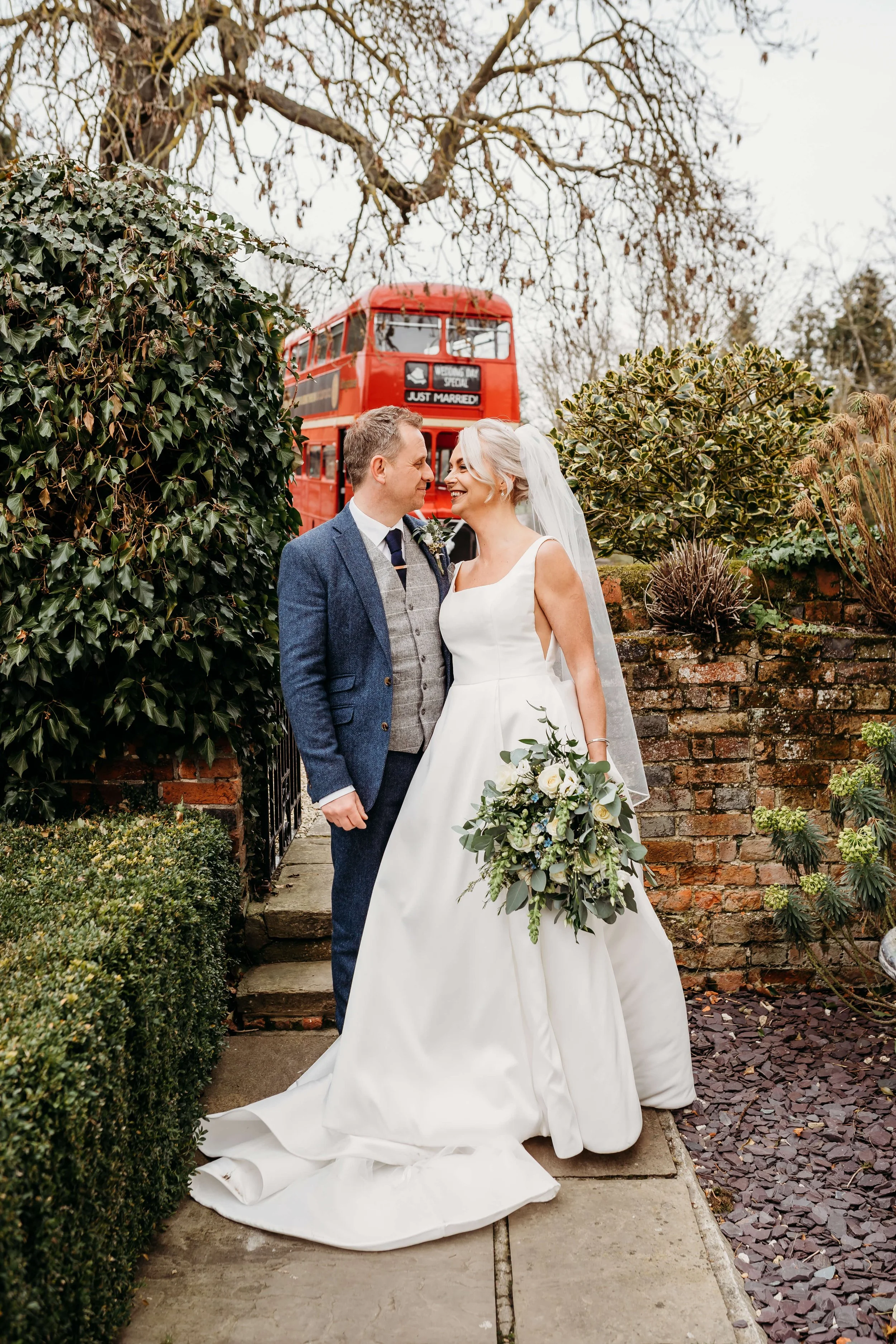 A bride and groom smile at each other outdoors on their wedding day, with a red double-decker bus in the background displaying the message "JUST MARRIED". The bride wears a white wedding gown and veil, holding a bouquet of white and green flowers. Th