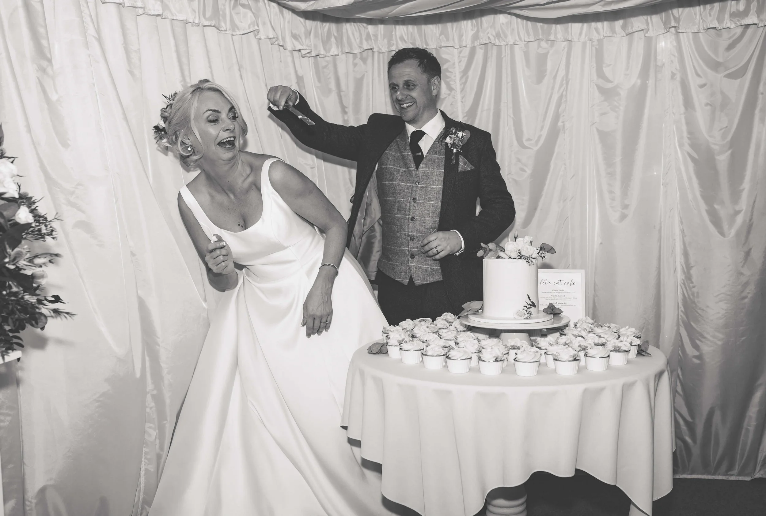 A bride and groom at their wedding reception cutting the cake, with cupcakes on the table, in black and white.