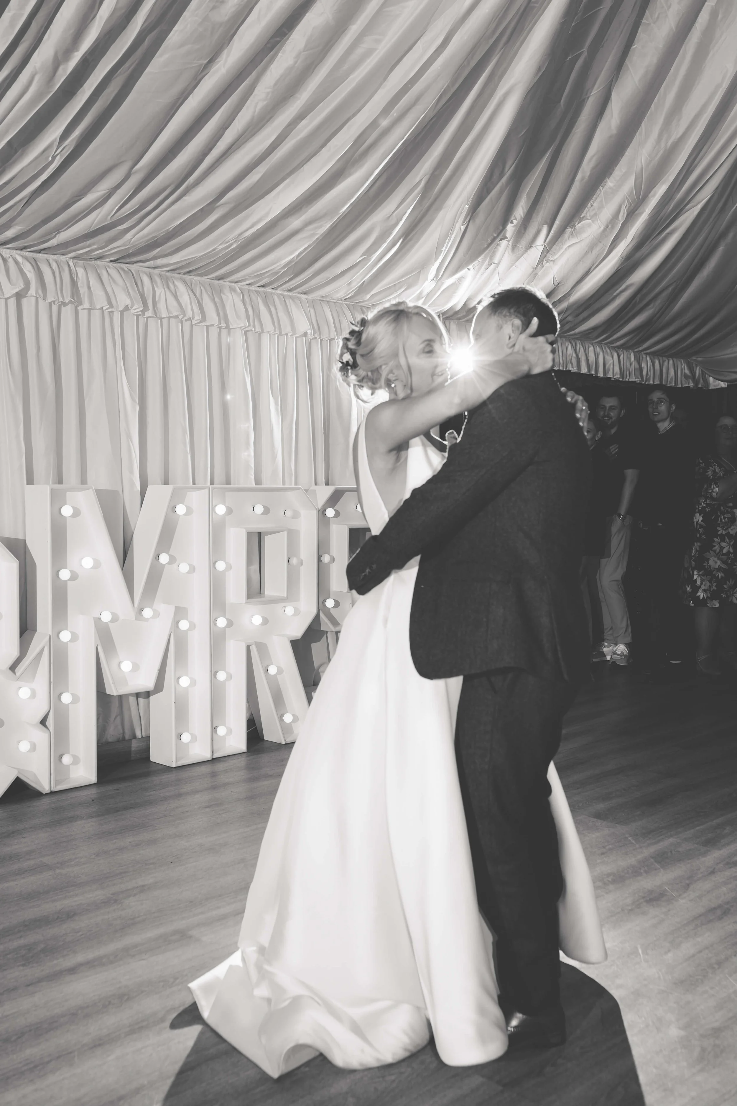 A bride and groom are dancing together at their wedding reception, with decorative large lit-up letters behind them.