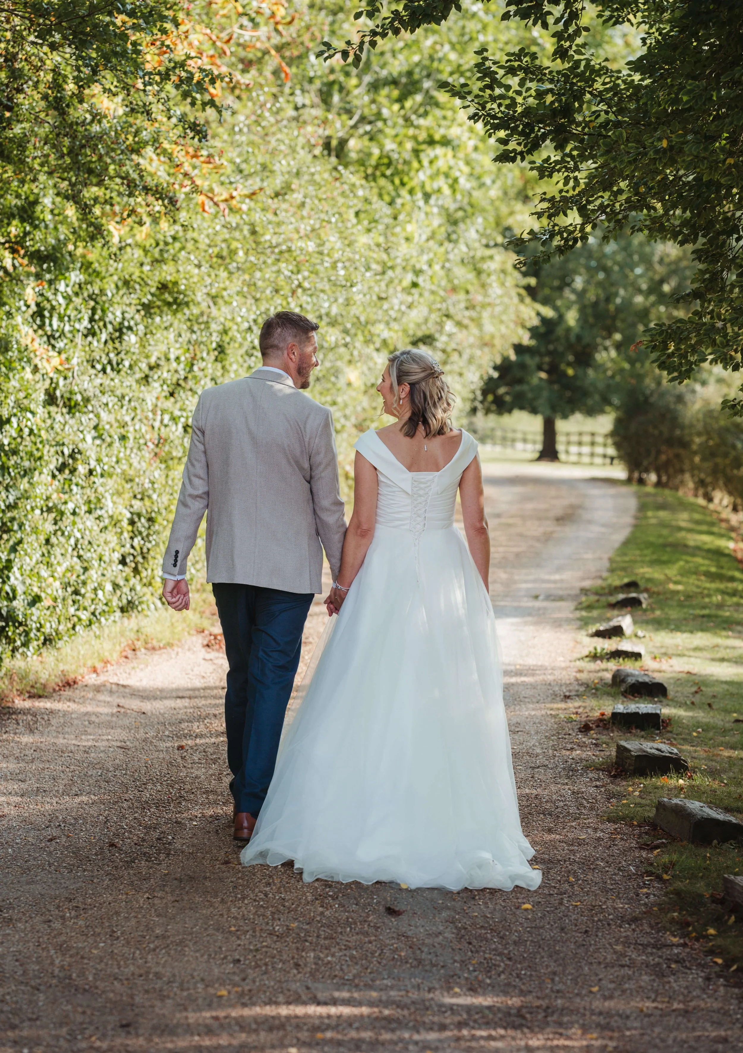 A newlywed couple walking hand-in-hand on a dirt path surrounded by green trees and foliage during daylight, with a wooden fence in the background.