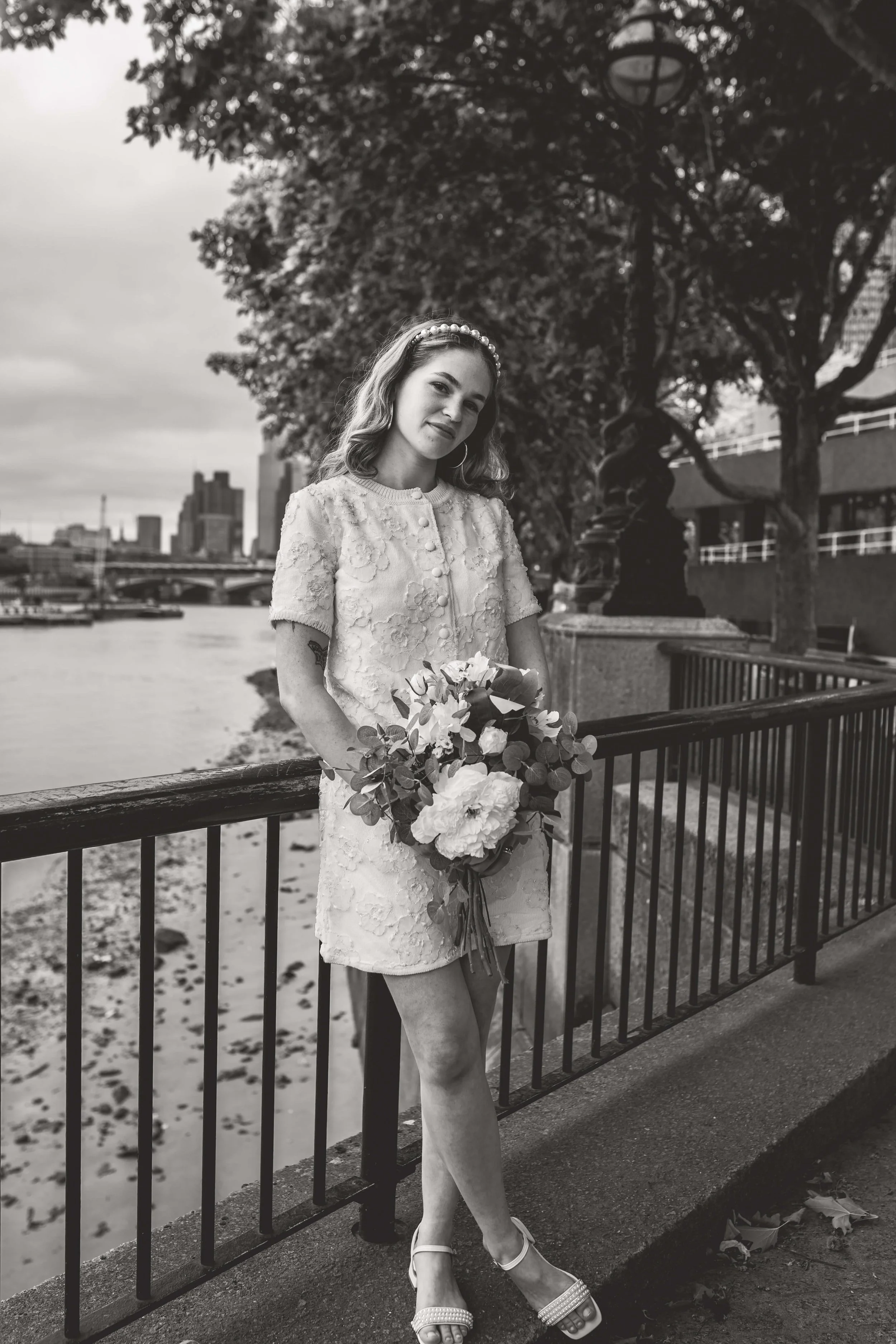 A woman in a short dress holding a bouquet of flowers, standing by a railing near a river with city buildings in the background, black and white photo, outdoors, possibly before or after a wedding.