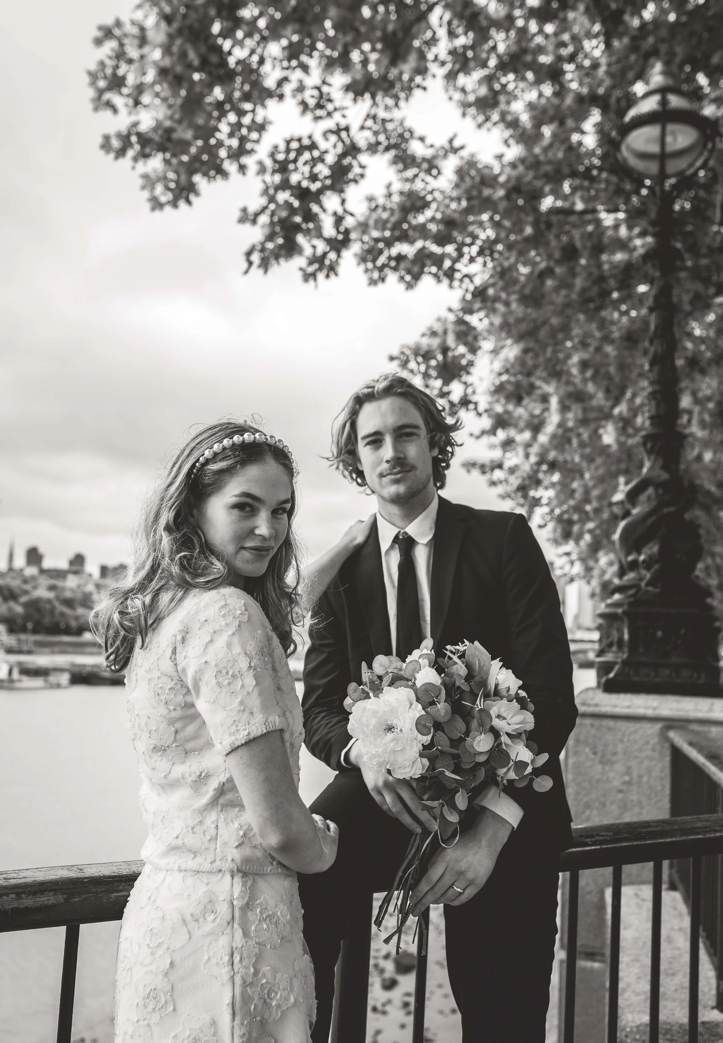 Black and white photo of a bride and groom standing outdoors near a river with city skyline in the background. The bride is wearing a lace dress and the groom is in a suit holding a bouquet of flowers. They are posing together under a tree with a dec