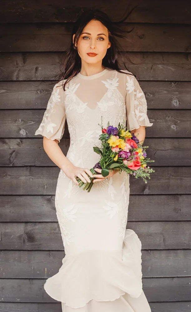 A woman with dark hair wearing a white wedding dress with lace details, holding a colorful bouquet of flowers, standing in front of a dark wooden wall.