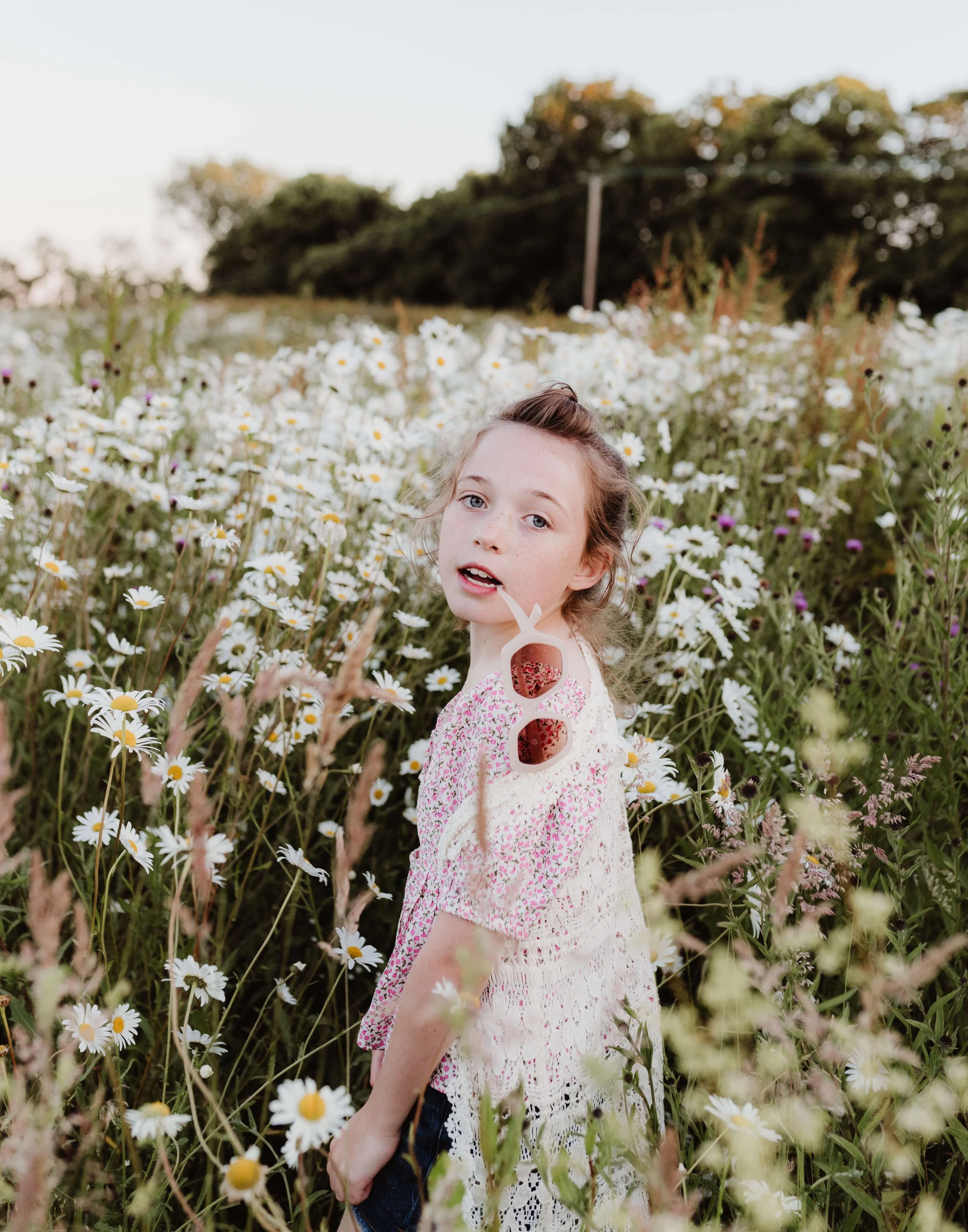 A young girl with curly brown hair and light skin standing in a field of daisies, wearing sunglasses on her shoulder and a white crochet top, with trees in the background and soft natural lighting.