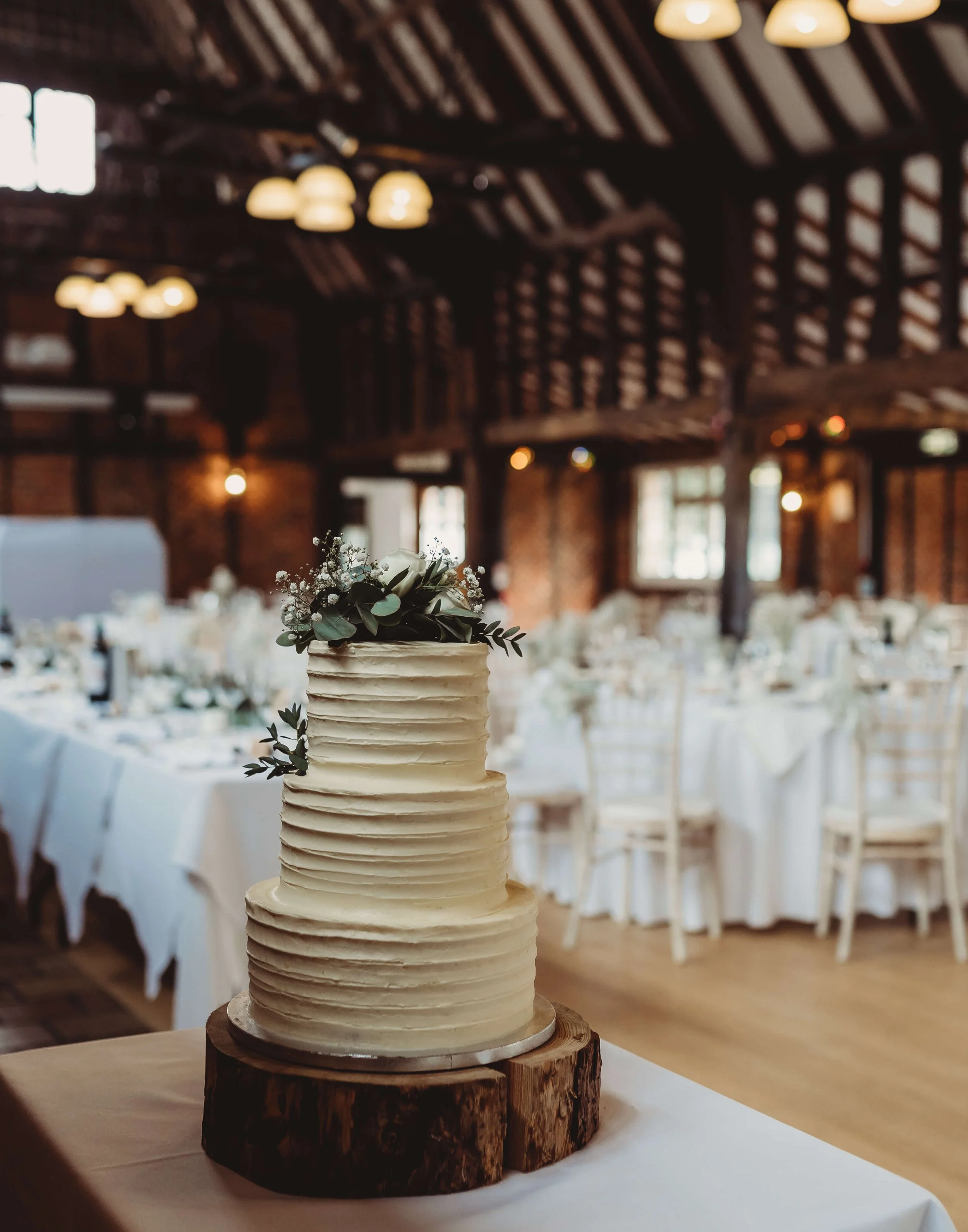 Elegant multi-tiered wedding cake with white frosting and floral decorations, placed on a wooden slab in a rustic barn wedding venue with tables and chairs in the background.