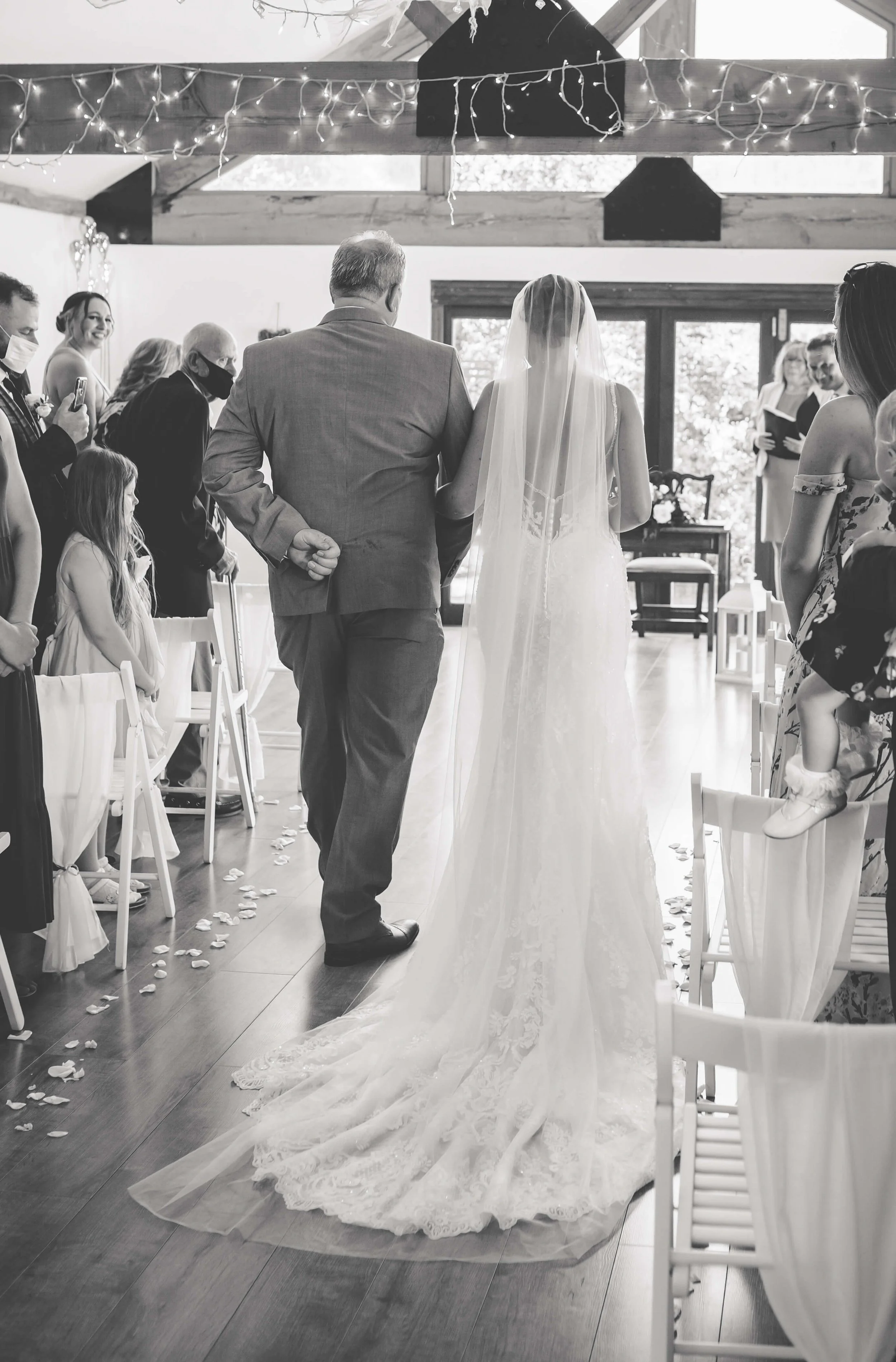 A bride walking down the aisle with an older man, possibly her father, at a wedding ceremony. Guests are seated on either side, some wearing masks, and there are string lights and curtains decorating the venue.