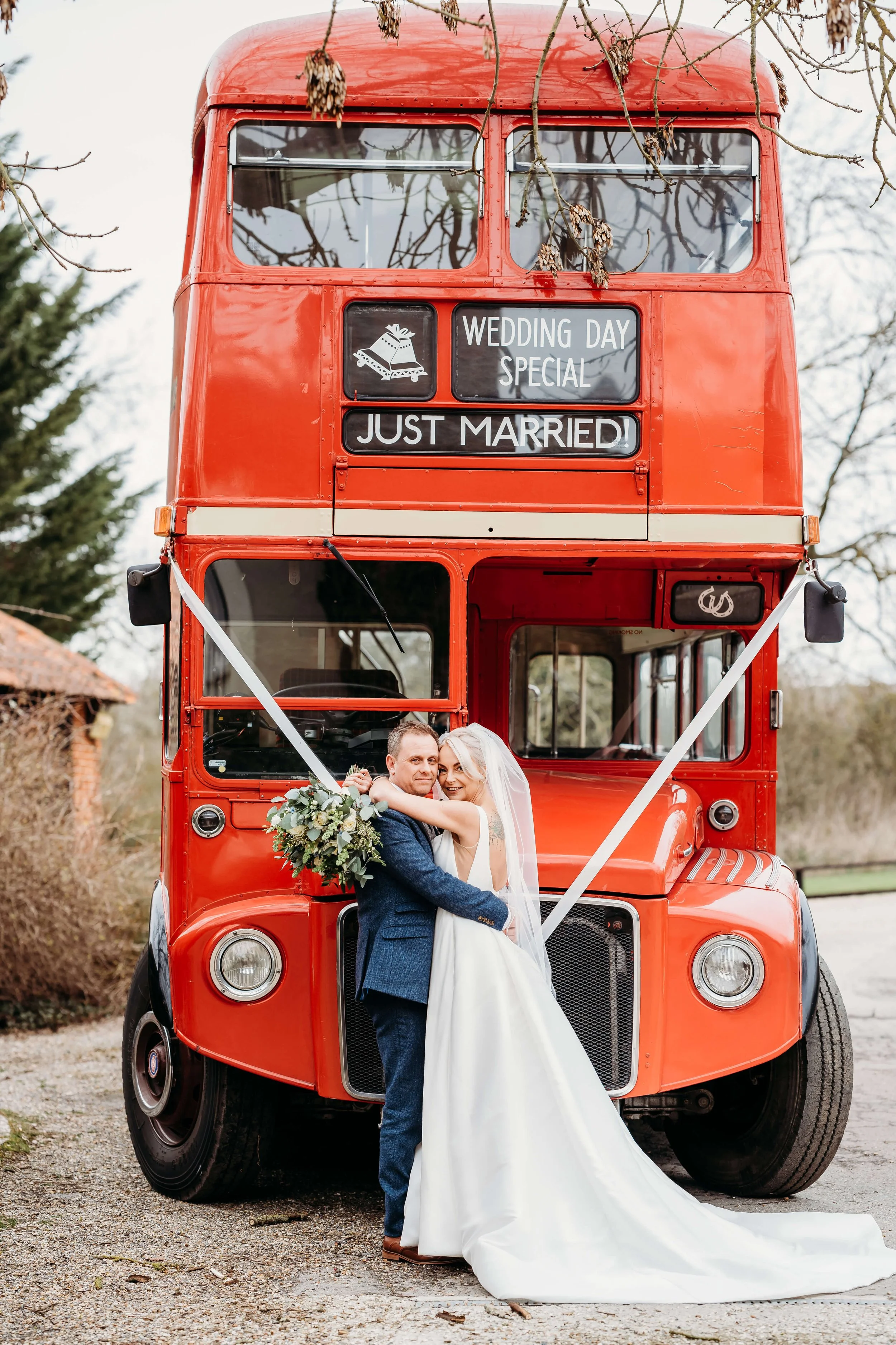 A newly married couple in wedding attire hugging in front of a vintage red double-decker bus decorated with signs that read 'Wedding Day Special' and 'Just Married.'
