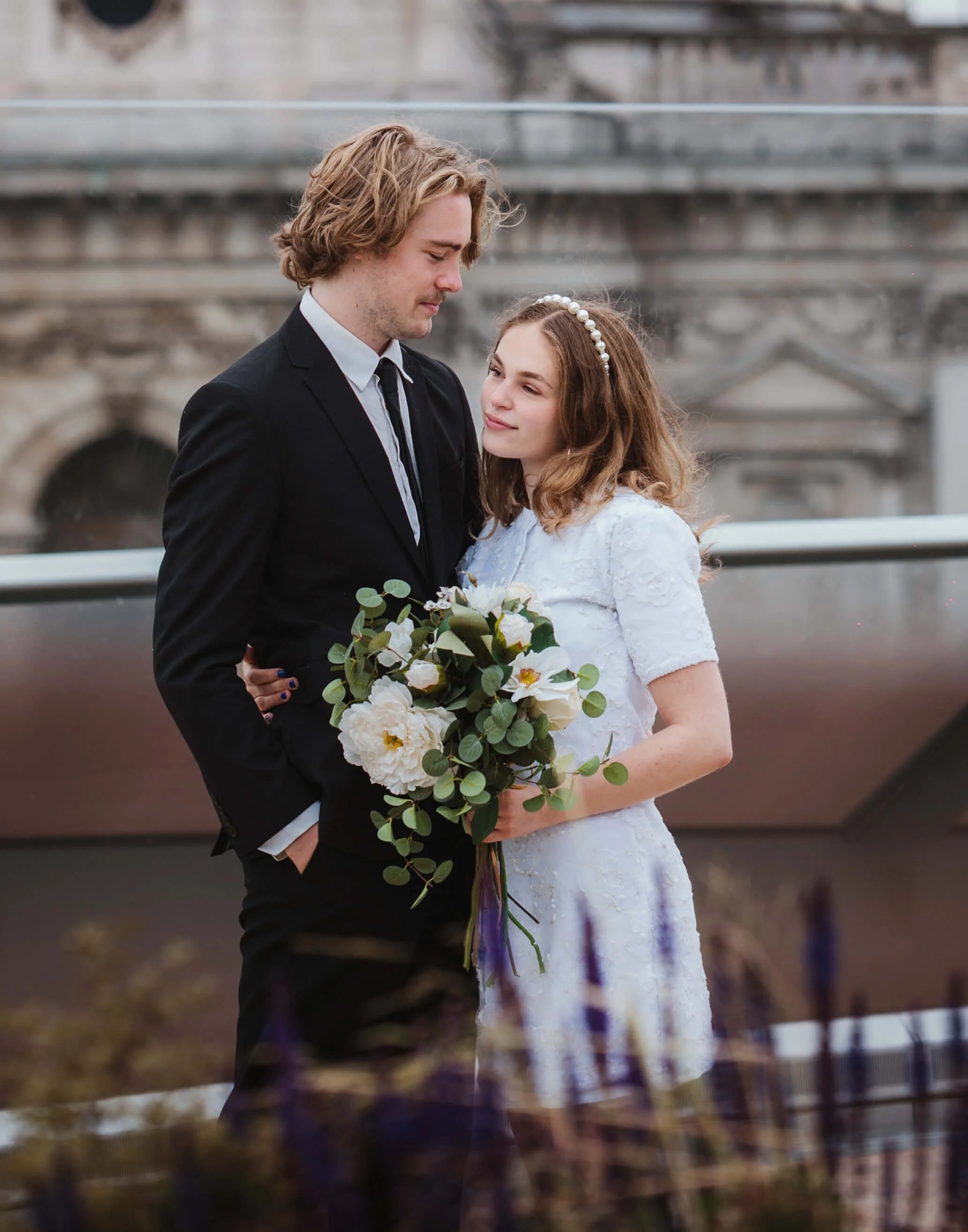 A young couple dressed in wedding attire standing close together outdoors, with the woman holding a bouquet of white flowers and greenery, and a historic building in the background.