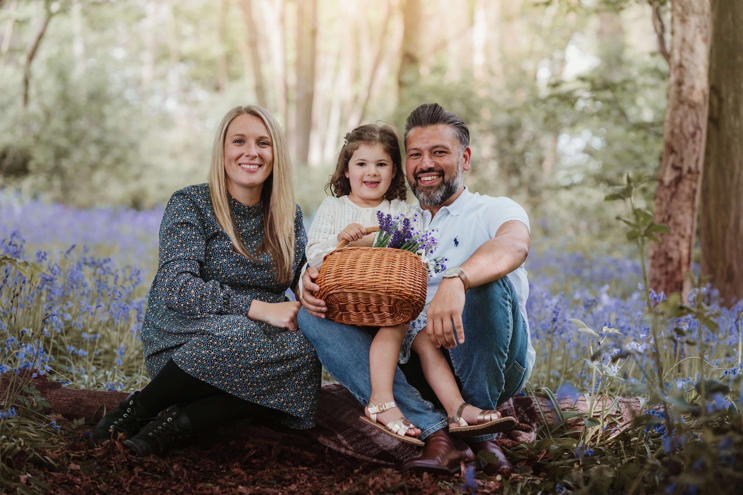 A family of three sitting on a blanket in a forest with blue flowers, smiling at the camera. The mother has long blonde hair, wearing a dark floral dress. The father has short dark hair and a beard, wearing a white polo shirt. The young girl with cur