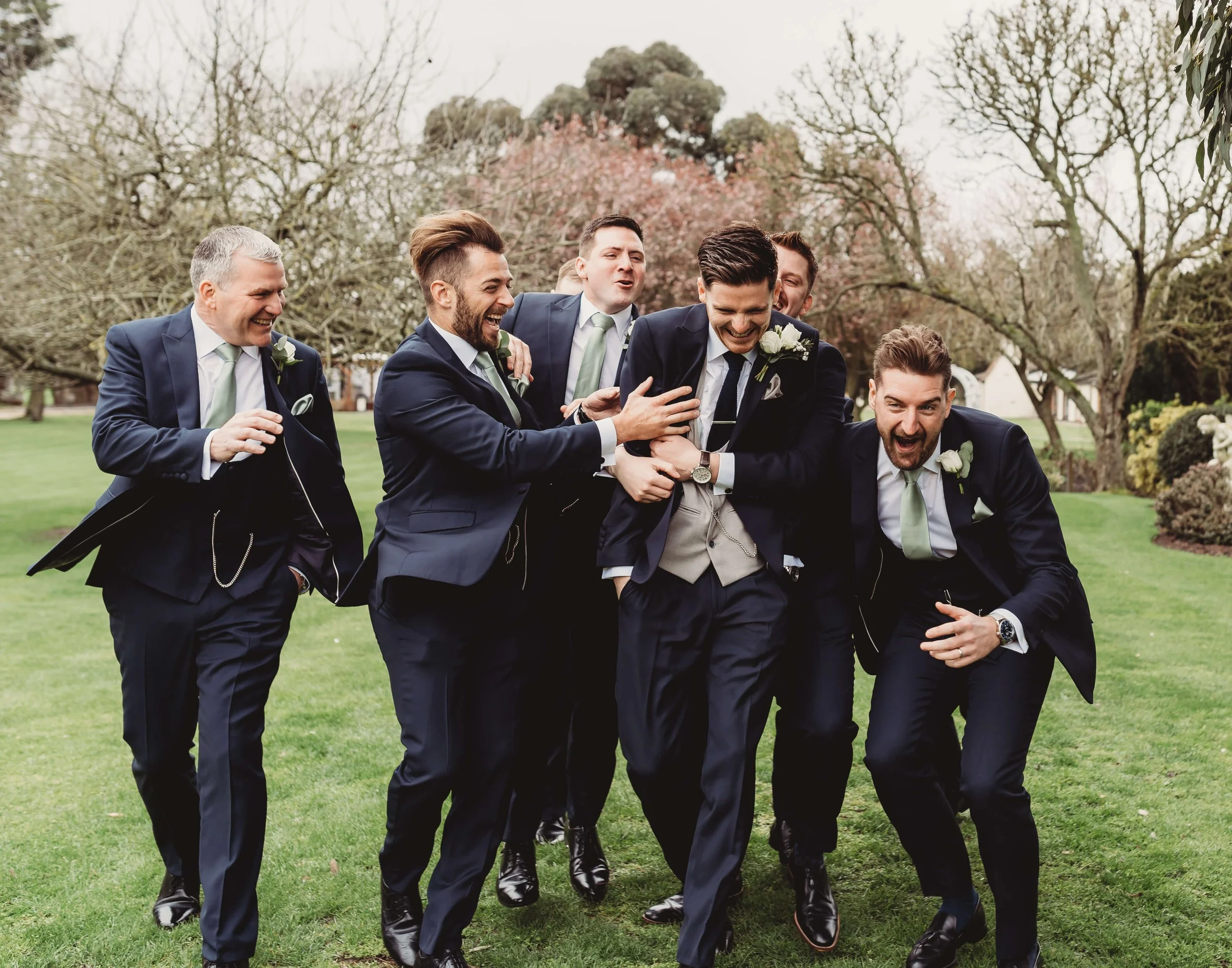Group of seven groomsmen in suits laughing and playing together outdoors on a grassy area, surrounded by trees at wedding.